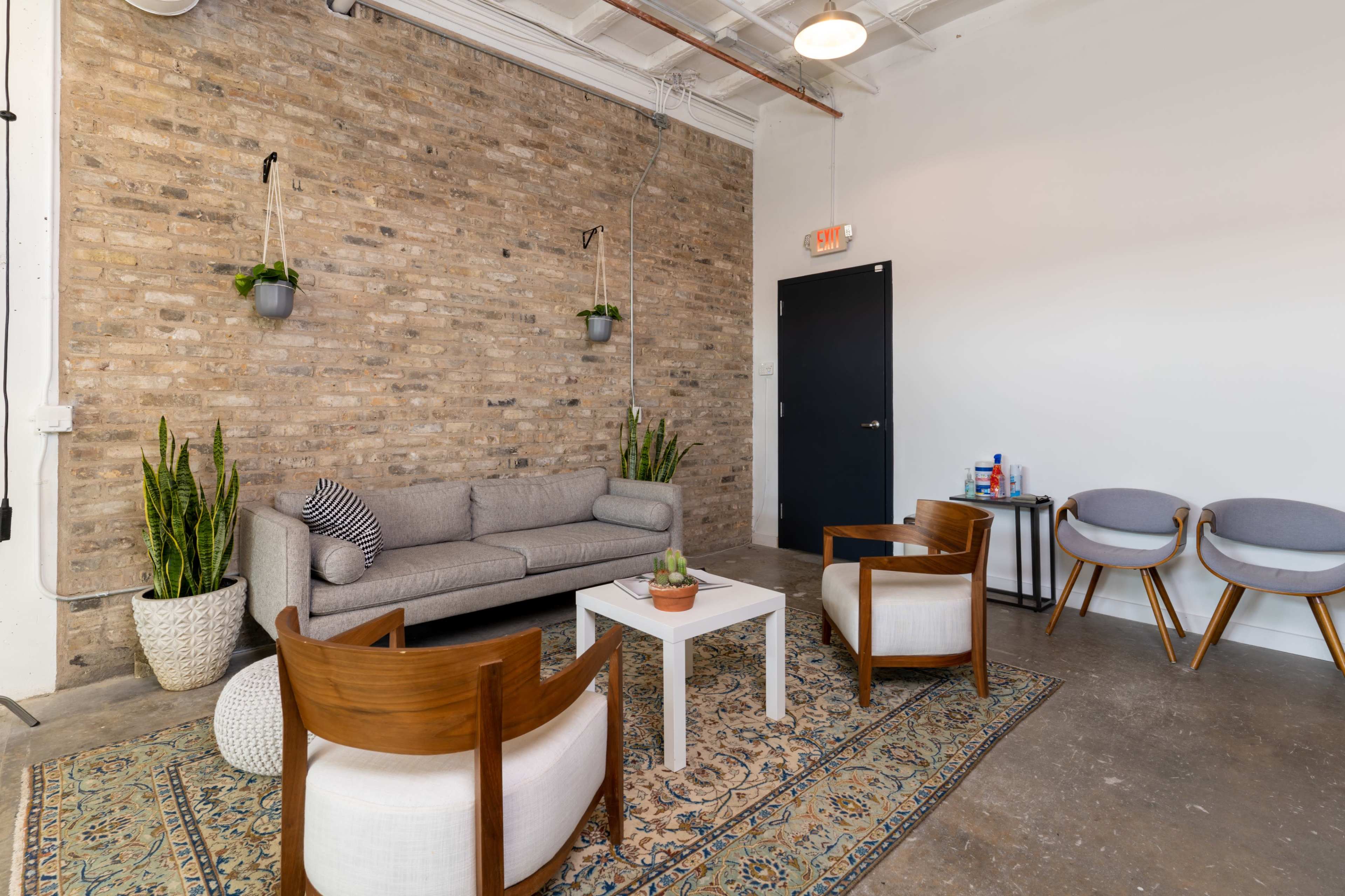 A seating area with a gray sofa, two wooden chairs, a coffee table, and a patterned rug against a brick wall with potted plants.