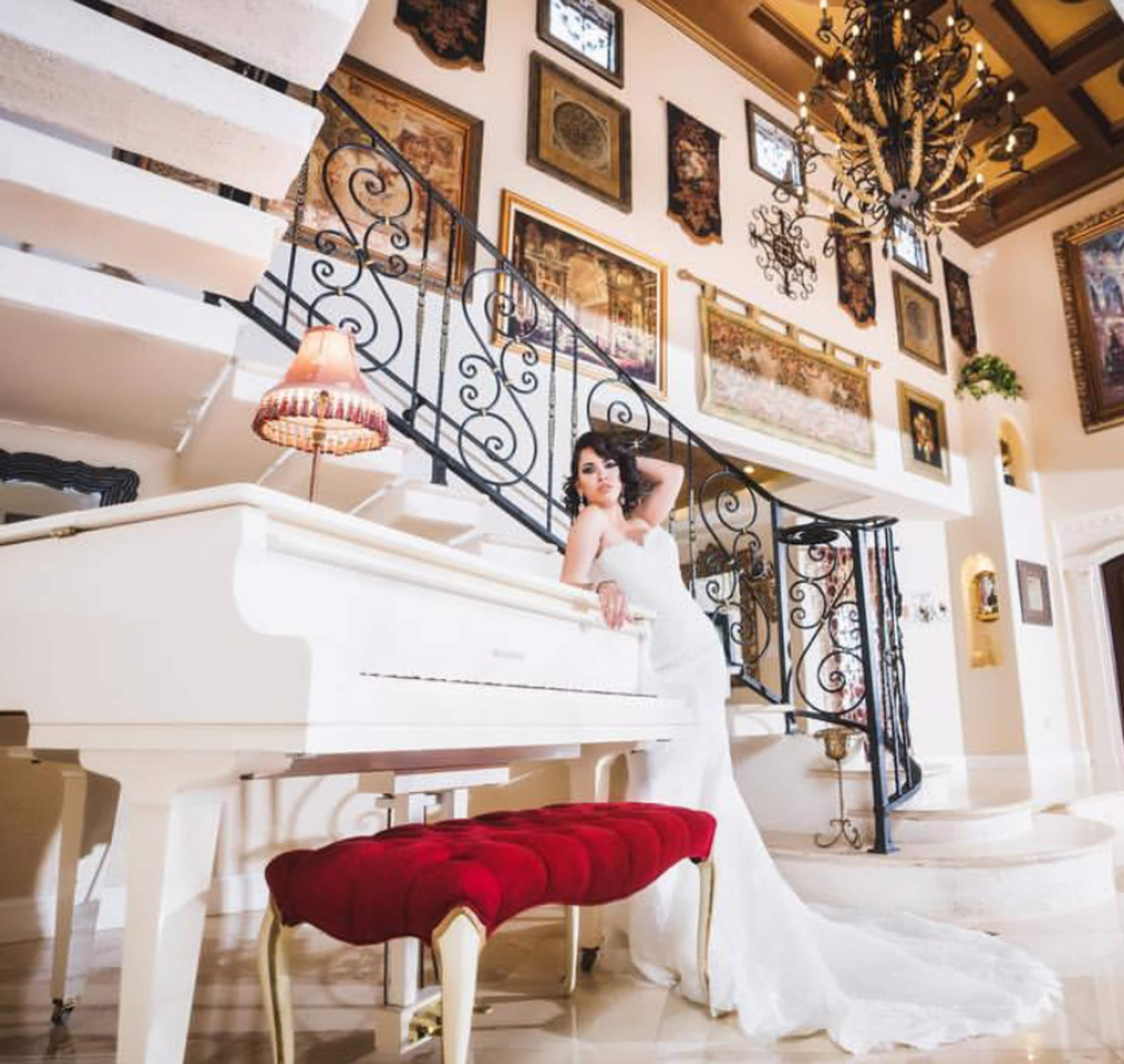 A woman in a white gown poses beside a grand white piano in a richly decorated room featuring ornate staircases and framed artwork on the walls.