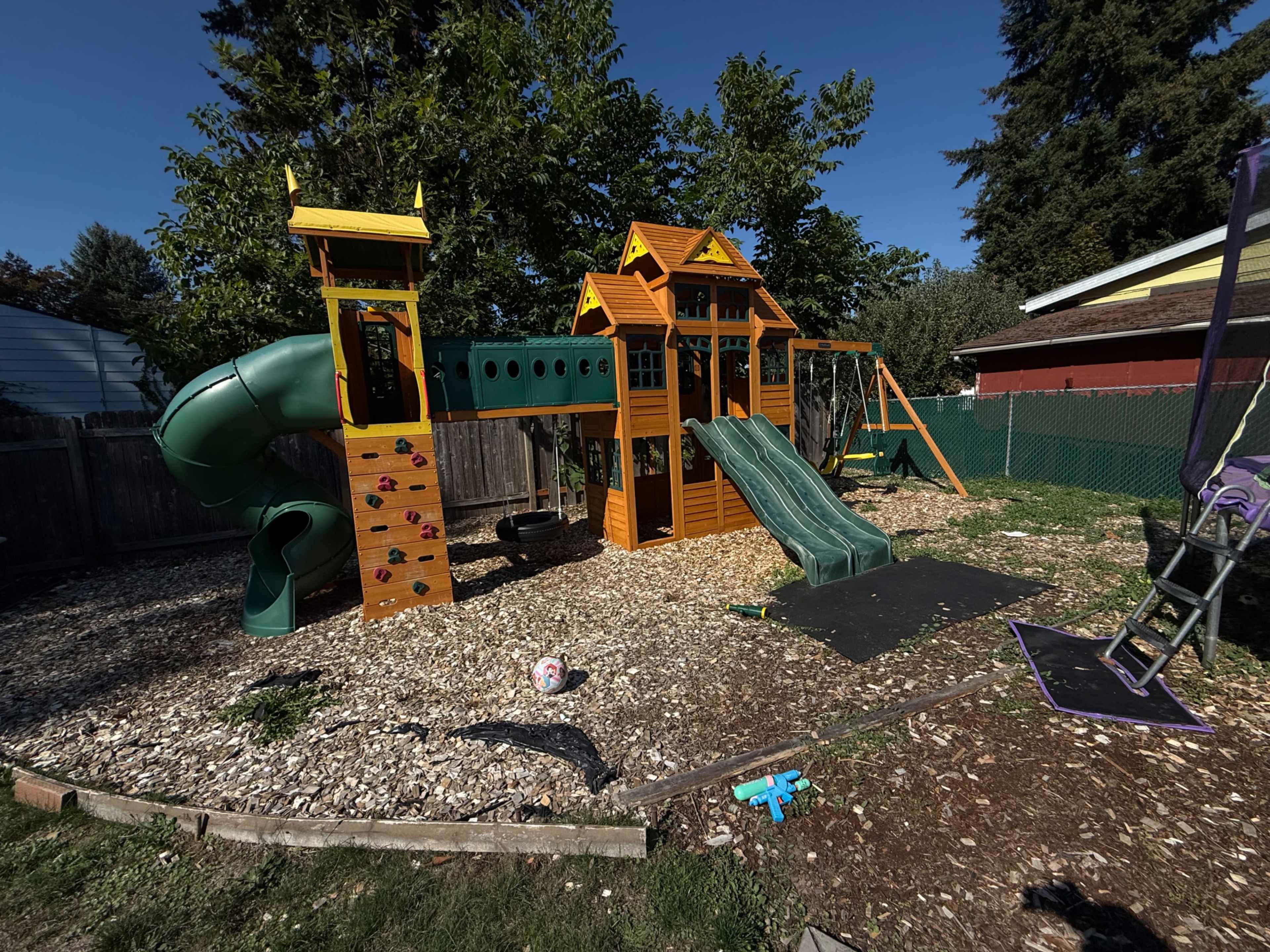 The image shows a brightly colored wooden playset in a backyard, featuring slides, swings, and climbing structures, surrounded by wood chips and grass.