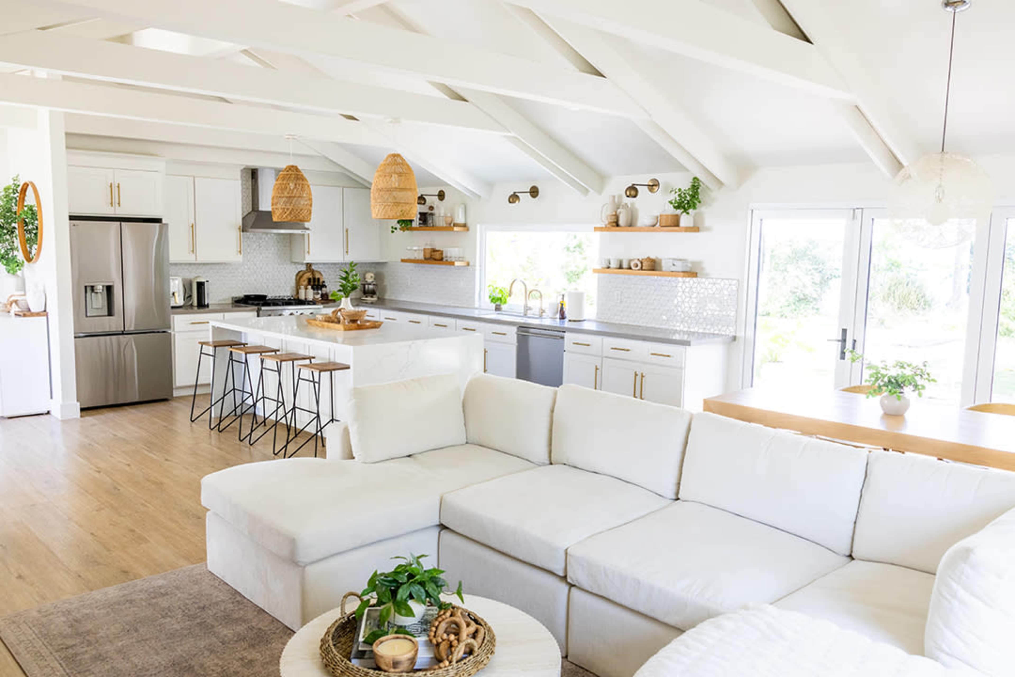 The image shows a modern, open-concept kitchen and living area with a large sectional sofa, a dining table, and natural light filtering through large windows.