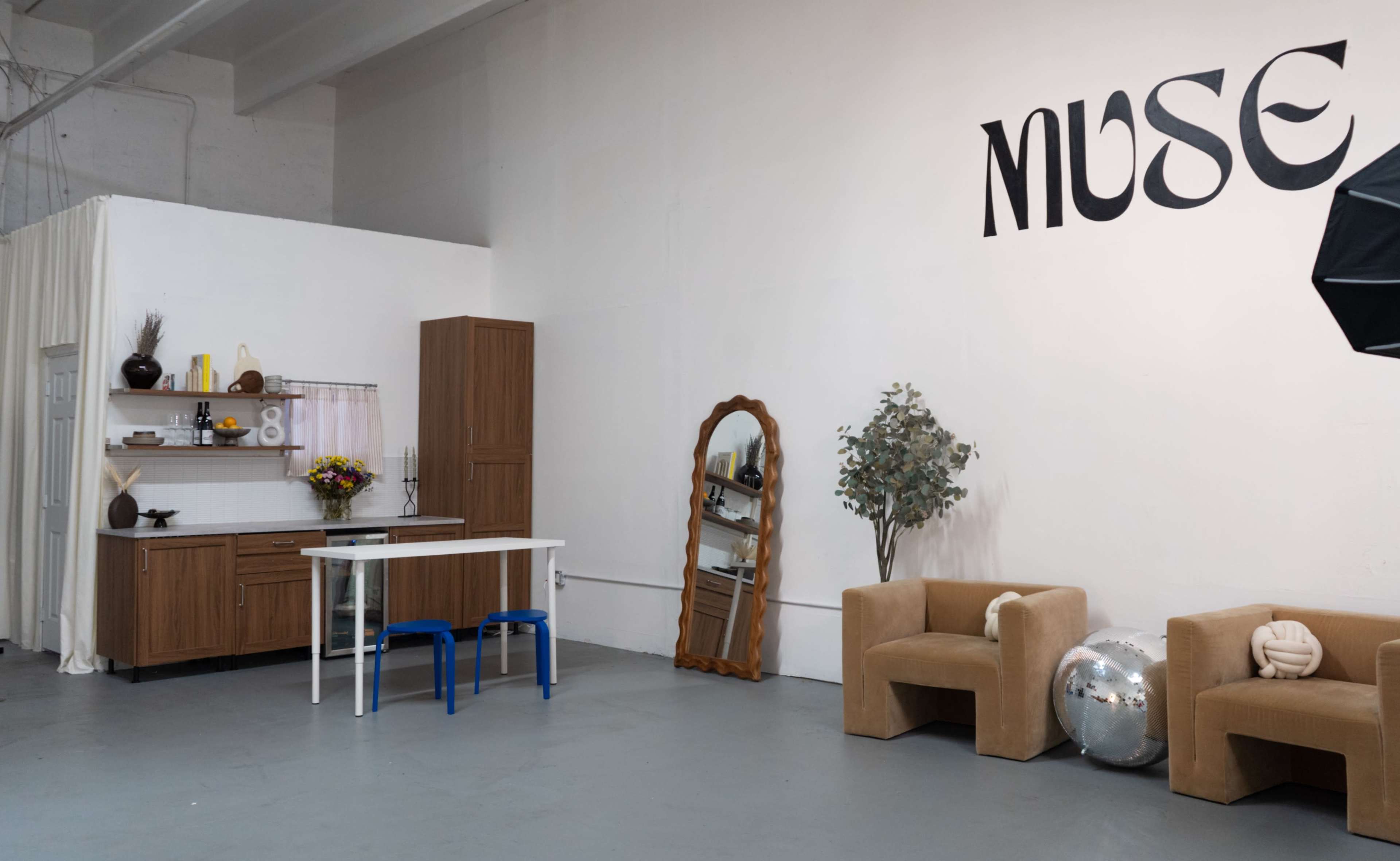 The image shows a minimalist studio space with a kitchenette, a mirror, a table with blue chairs, two brown chairs, and decorative items.