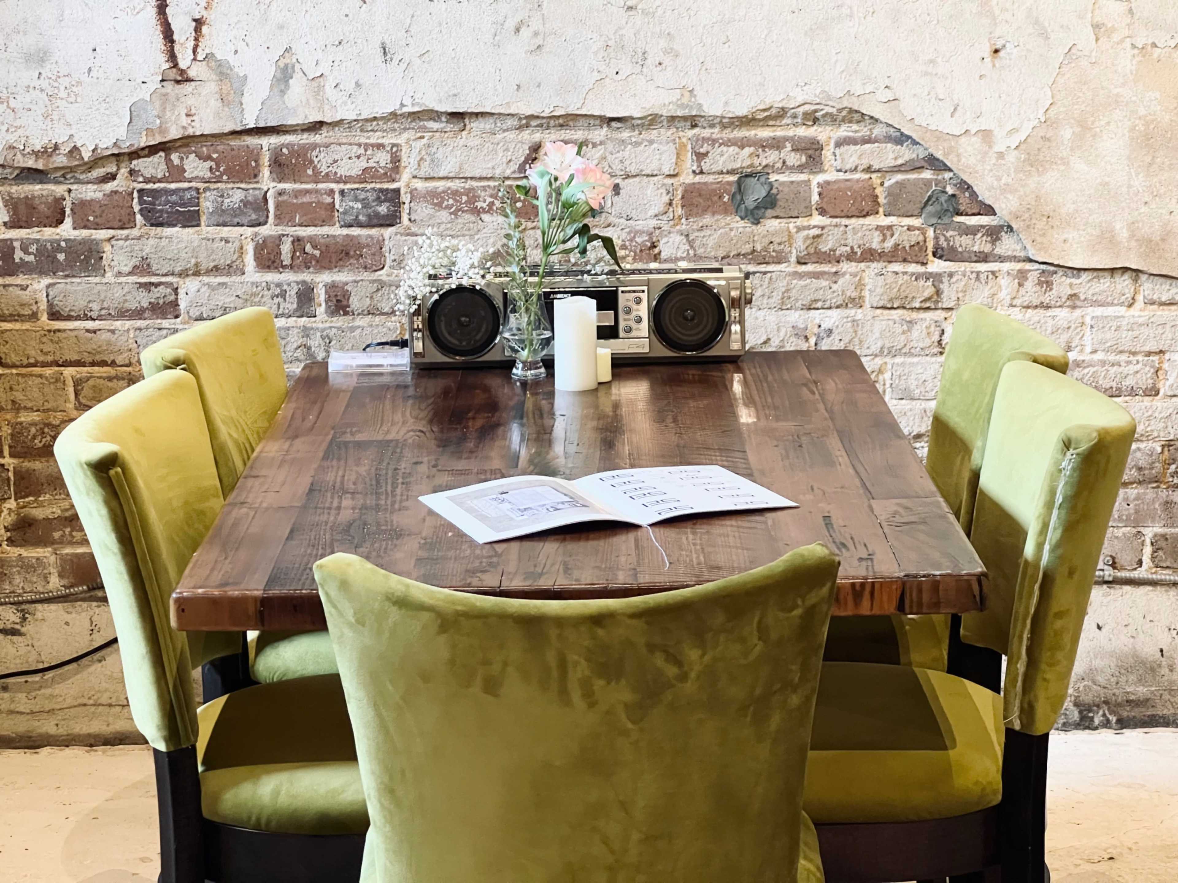 A wooden dining table surrounded by four green chairs, with a vintage boombox and a vase of flowers in the background against a brick wall.