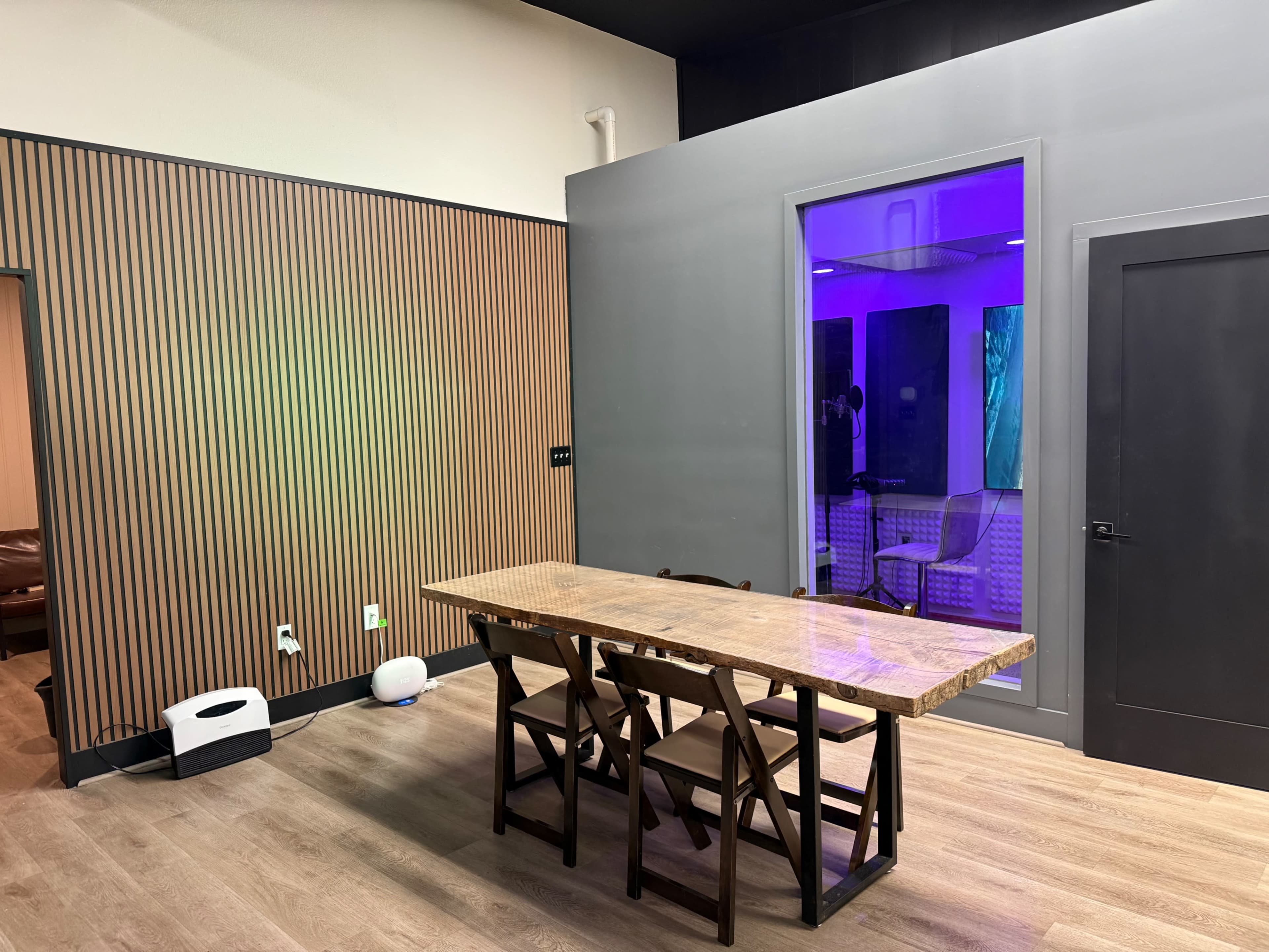 The image shows a modern interior space featuring a wooden table with chairs, a wall with vertical wooden panels, and a glass-enclosed area illuminated with purple light.