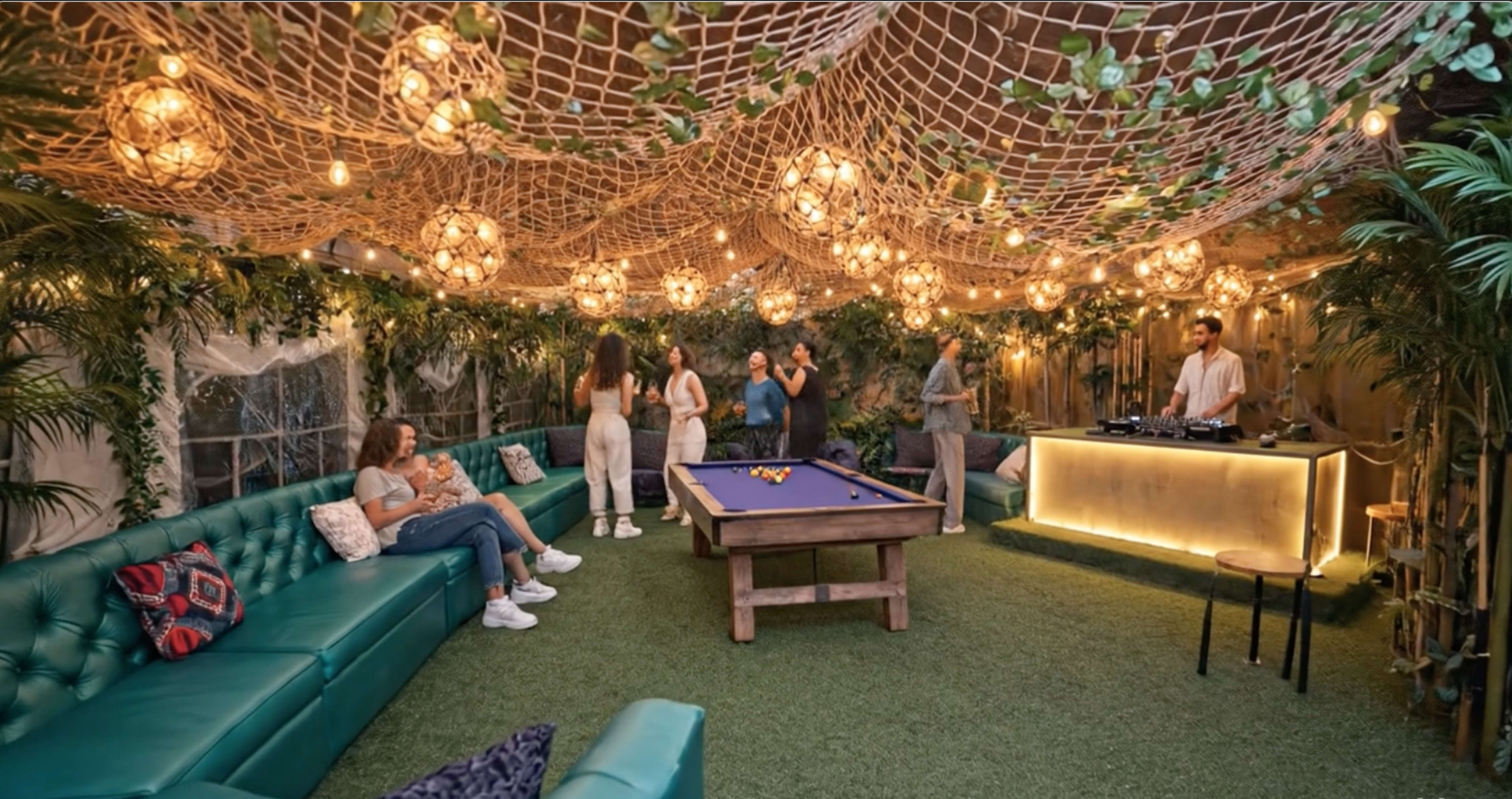 The image shows a vibrant indoor lounge area with a pool table, green sofas, and decorative string lights surrounded by plants.