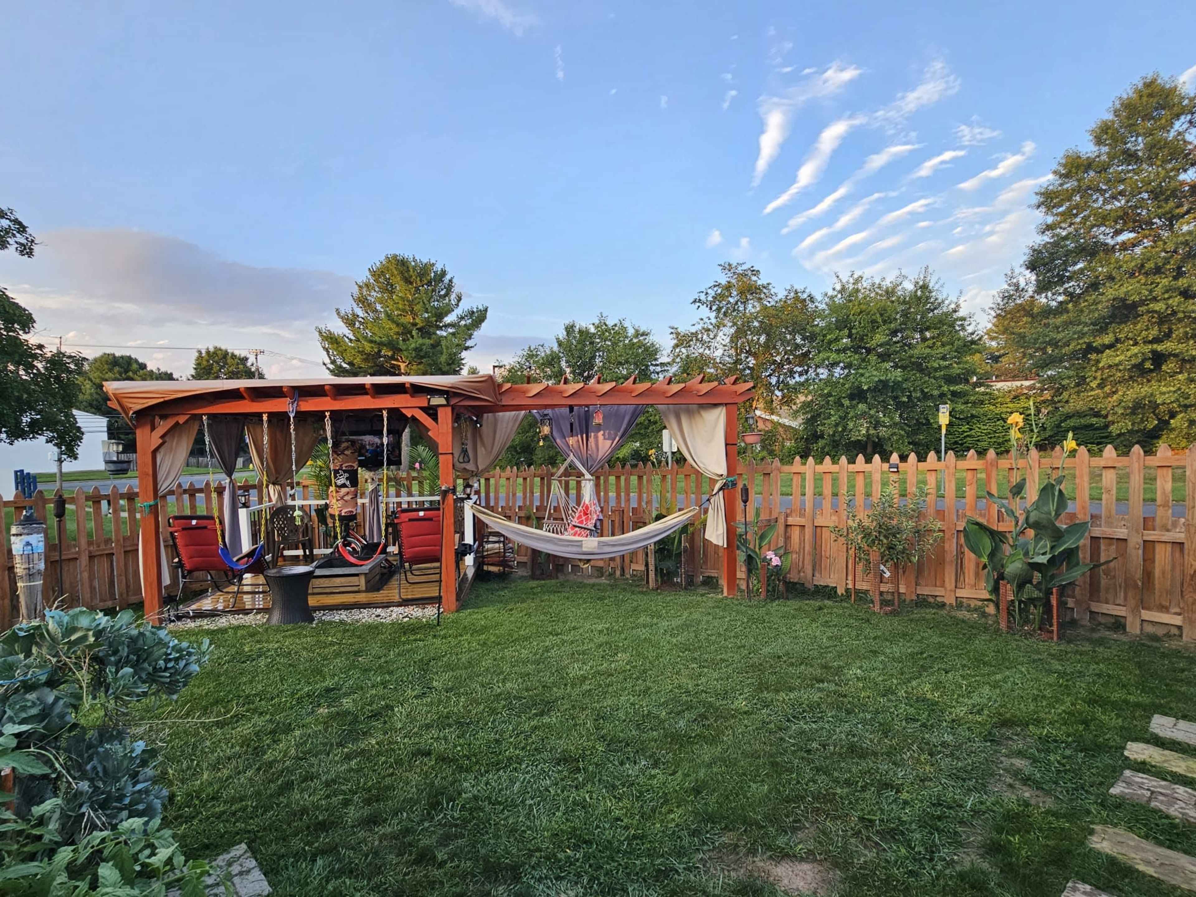 A backyard with a wooden pergola, two hammocks, comfortable seating, and a grassy area surrounded by a fence and various plants.