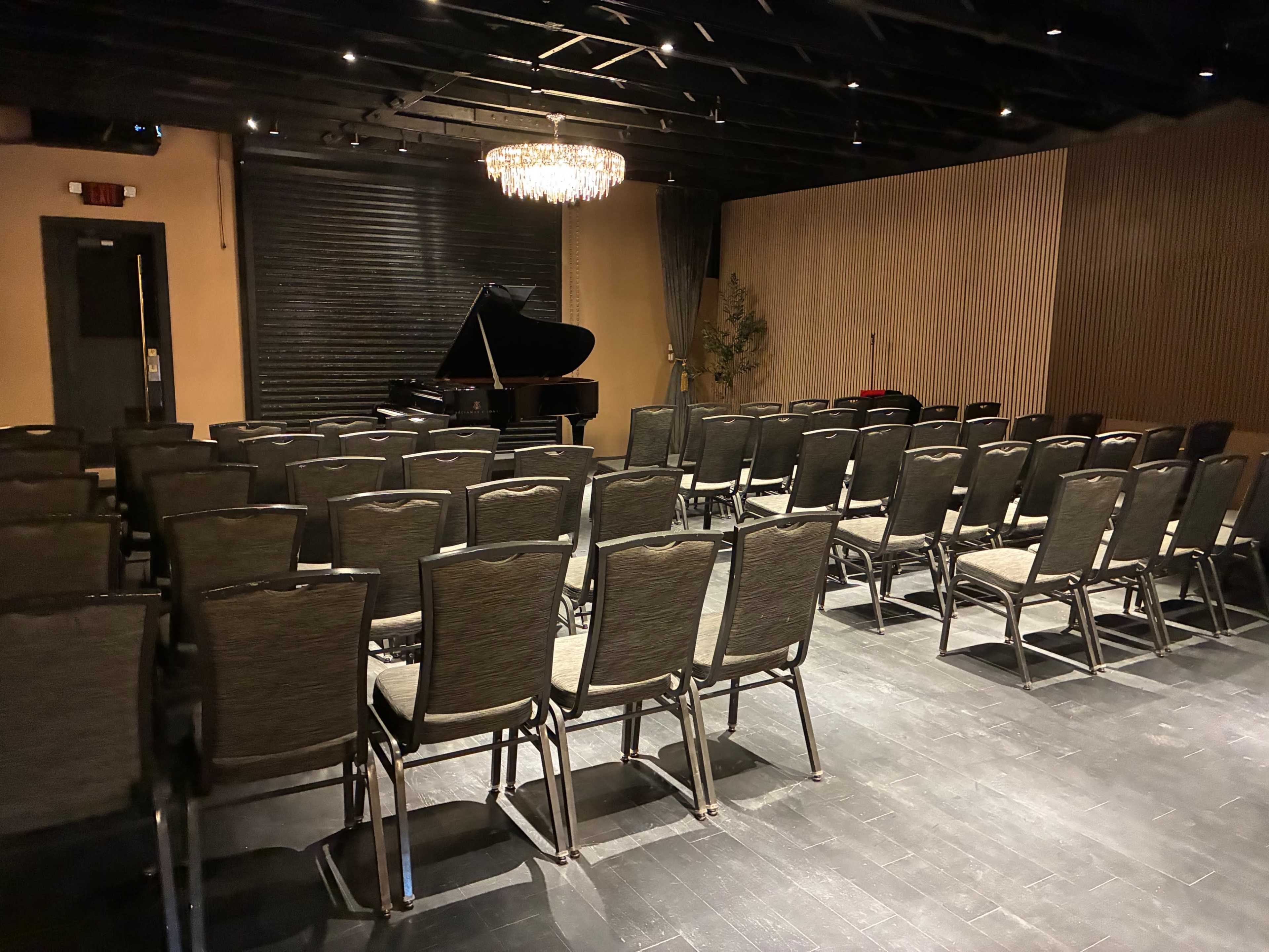 The image shows a nearly empty performance space with rows of black chairs arranged facing a grand piano under a chandelier.