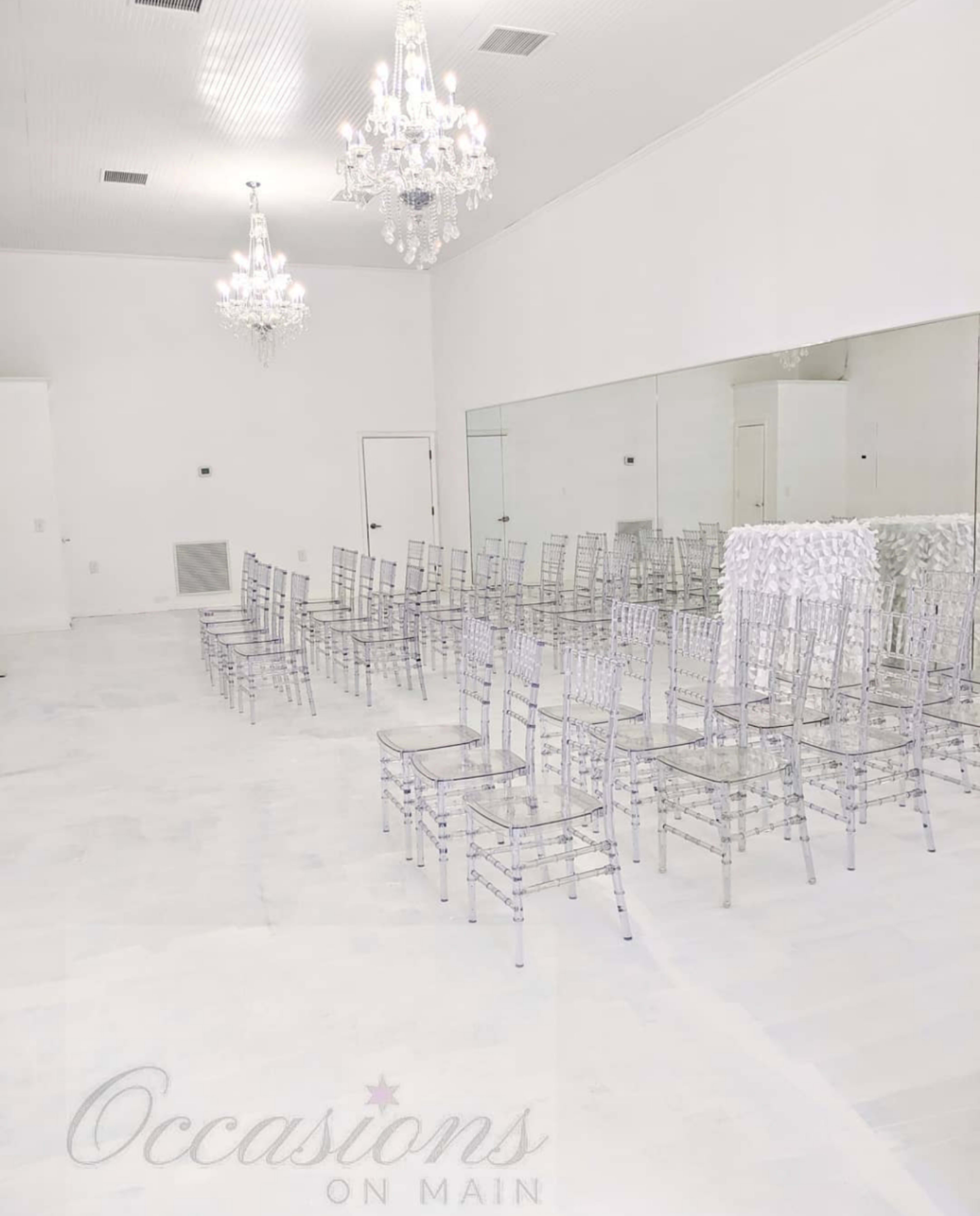 A bright, empty room features rows of transparent chairs and two chandeliers hanging from the ceiling, with a large mirror on one wall.