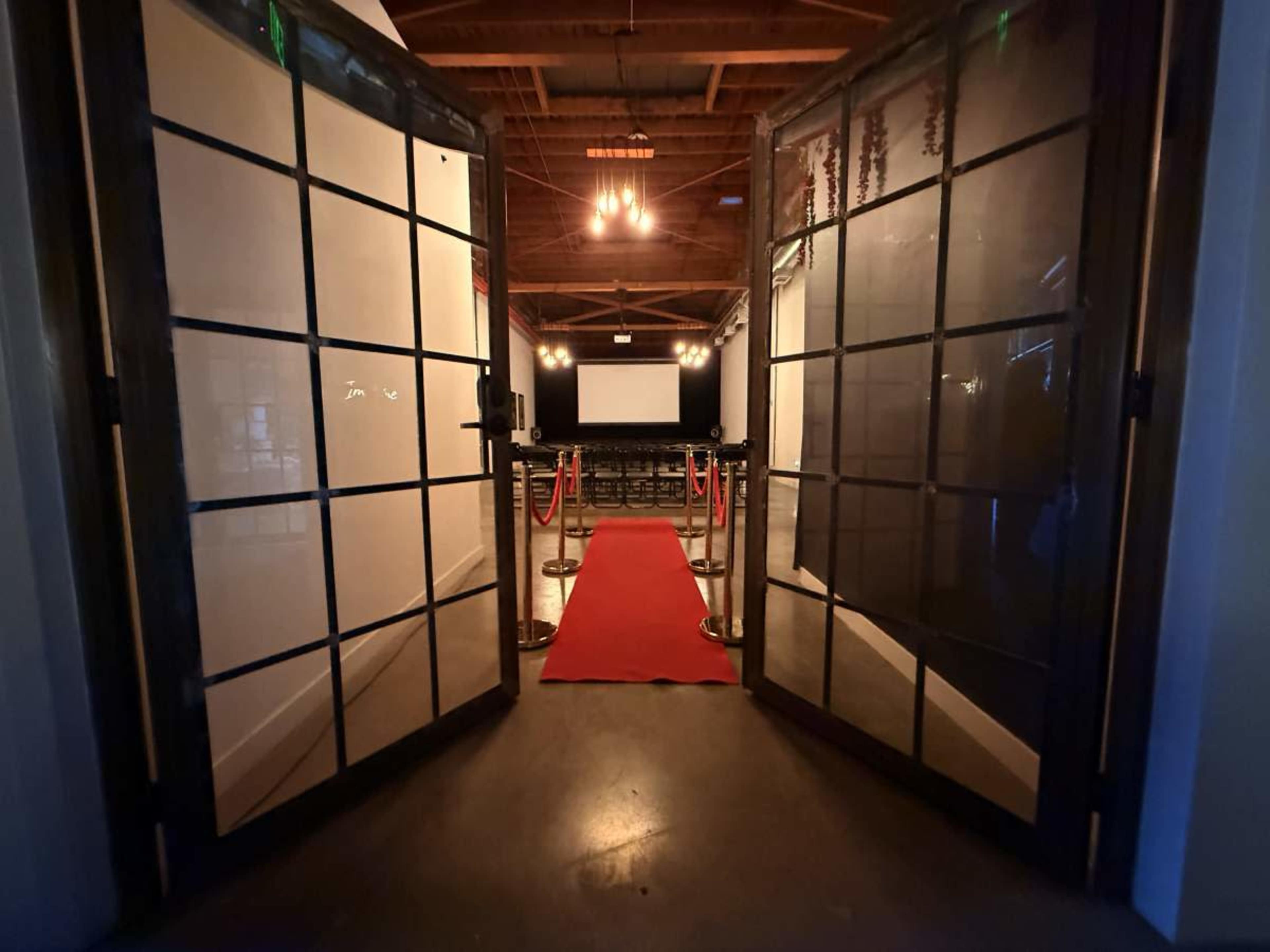 The image shows an entrance with double glass doors opening to a room featuring a red carpet and a projector screen at the far end.