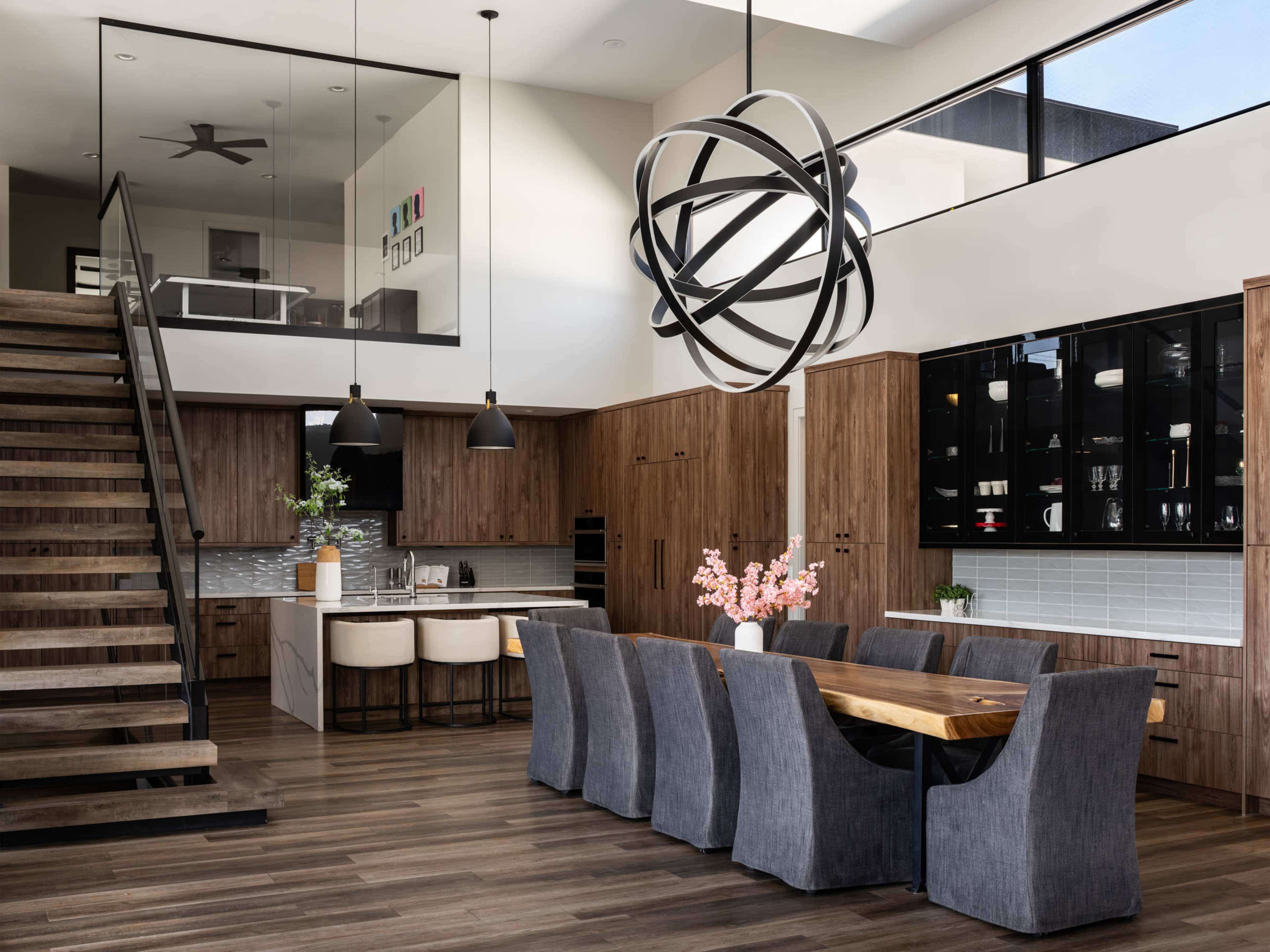 The image shows a modern kitchen and dining area with wooden cabinetry, a large dining table, and contemporary light fixtures.