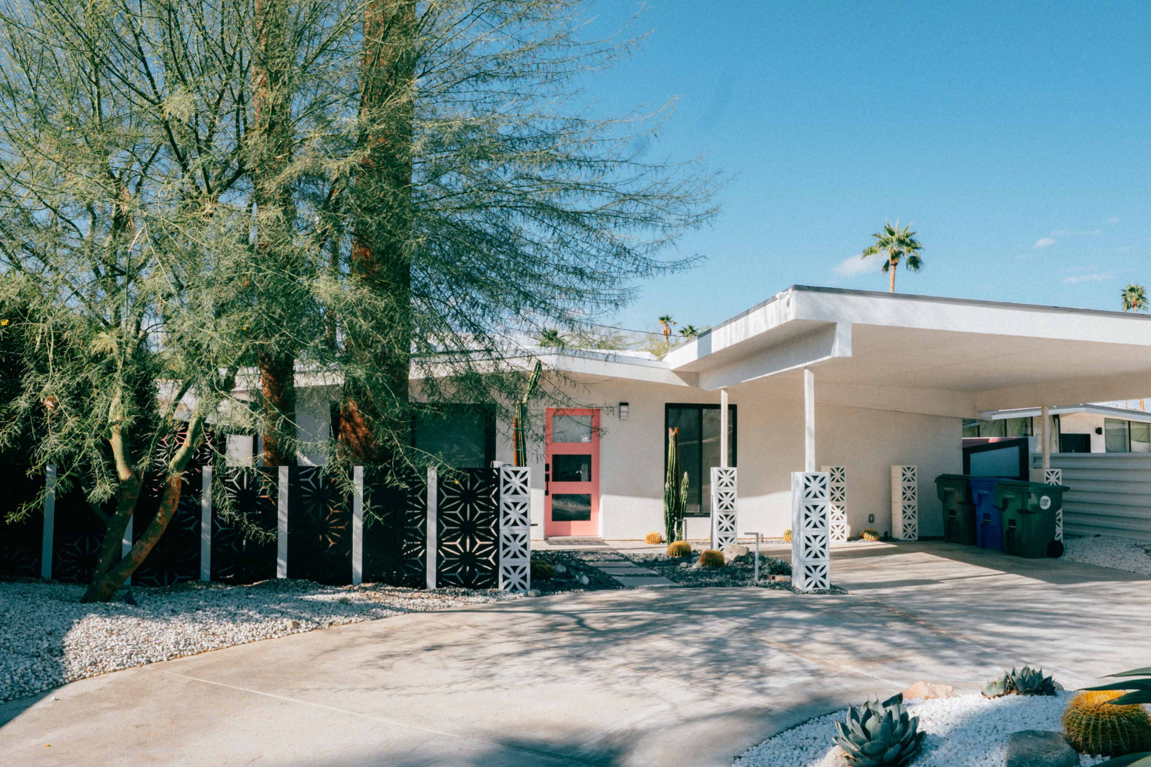 The image shows a modern single-story home with a unique architectural design, surrounded by desert landscaping and decorative concrete elements.
