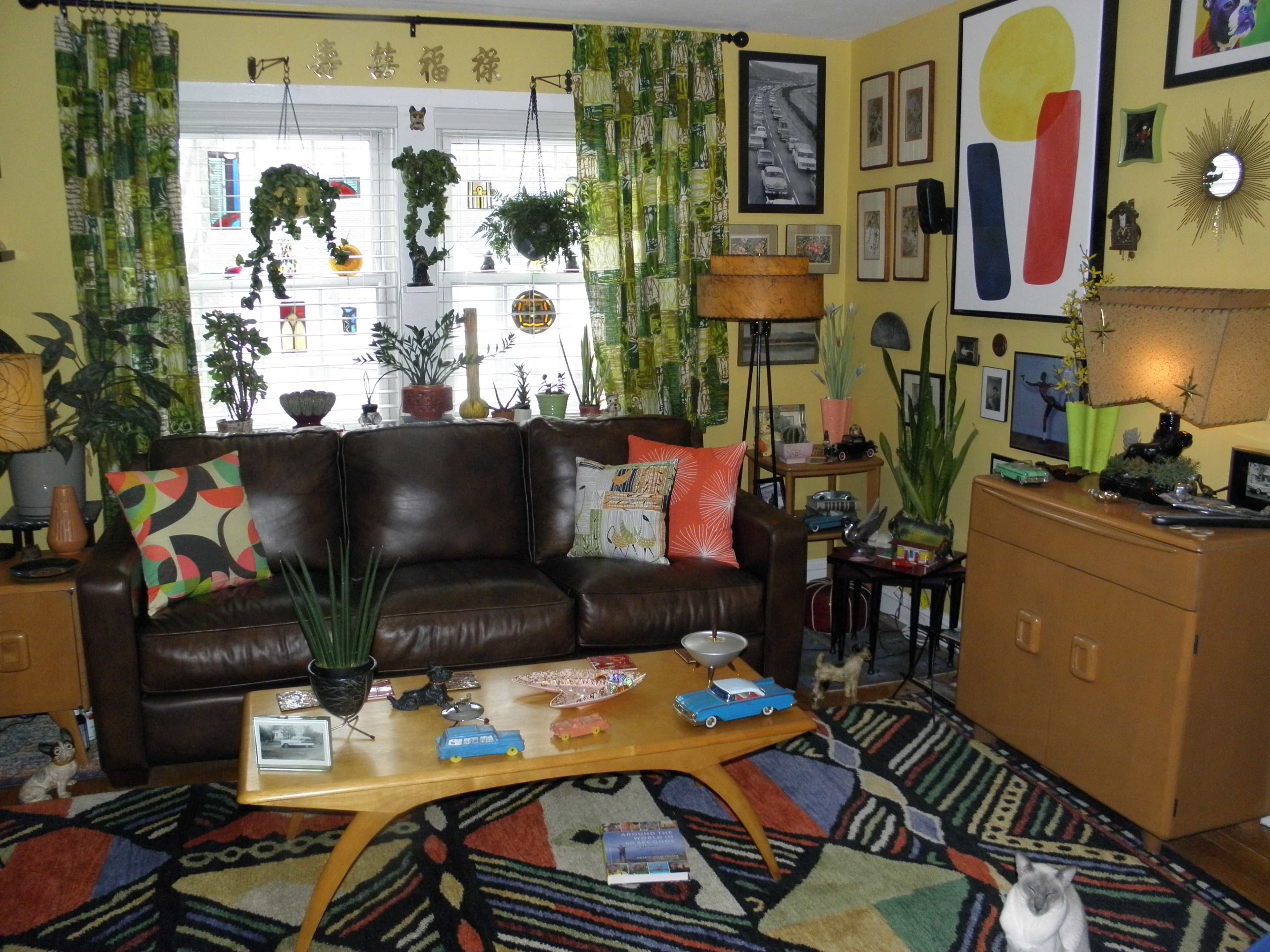 A living room features a brown leather sofa, a colorful area rug, plants, and various artwork on the walls.