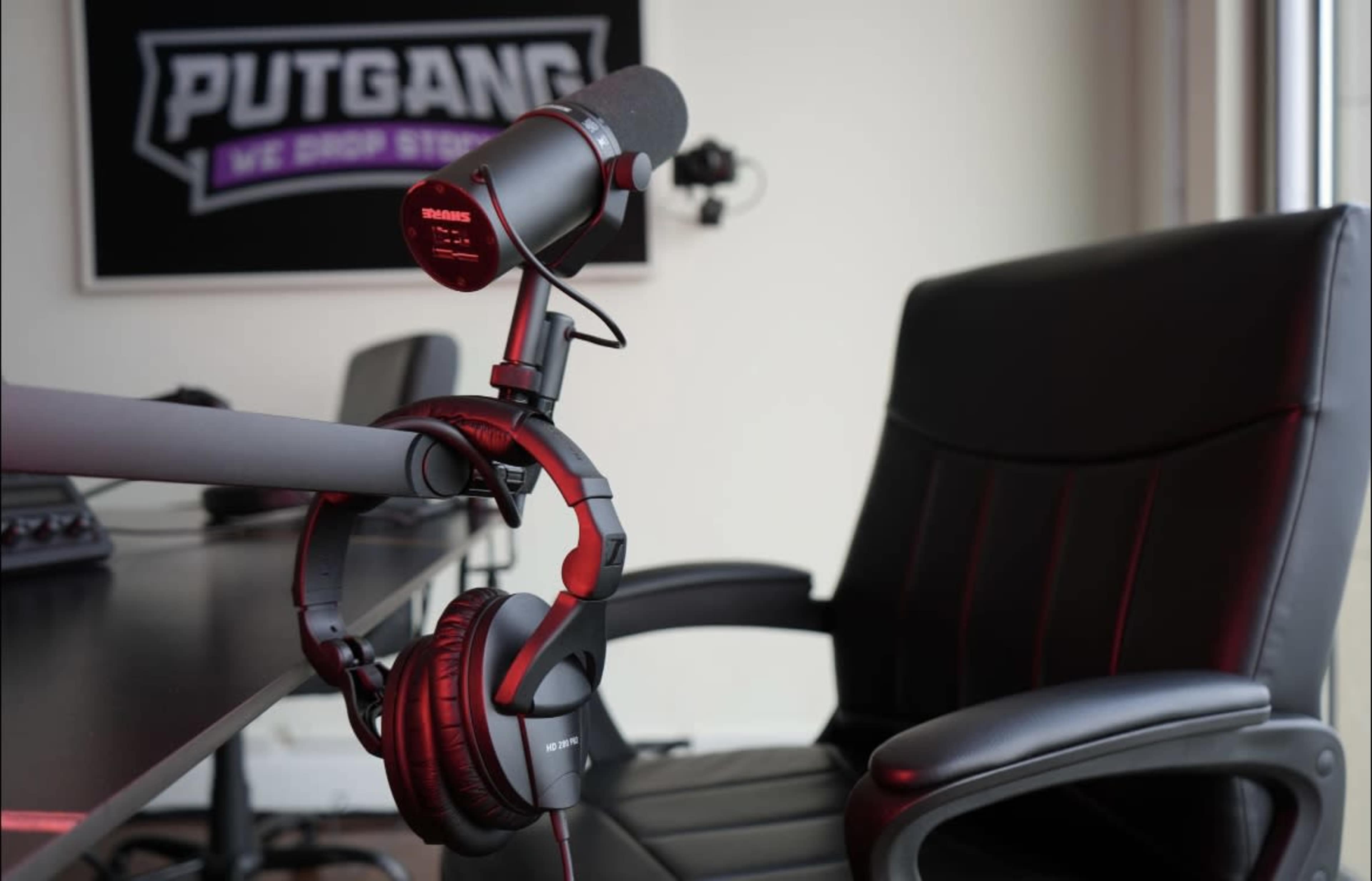 A microphone is mounted on a desk next to a pair of headphones, with a gaming chair visible in the background.