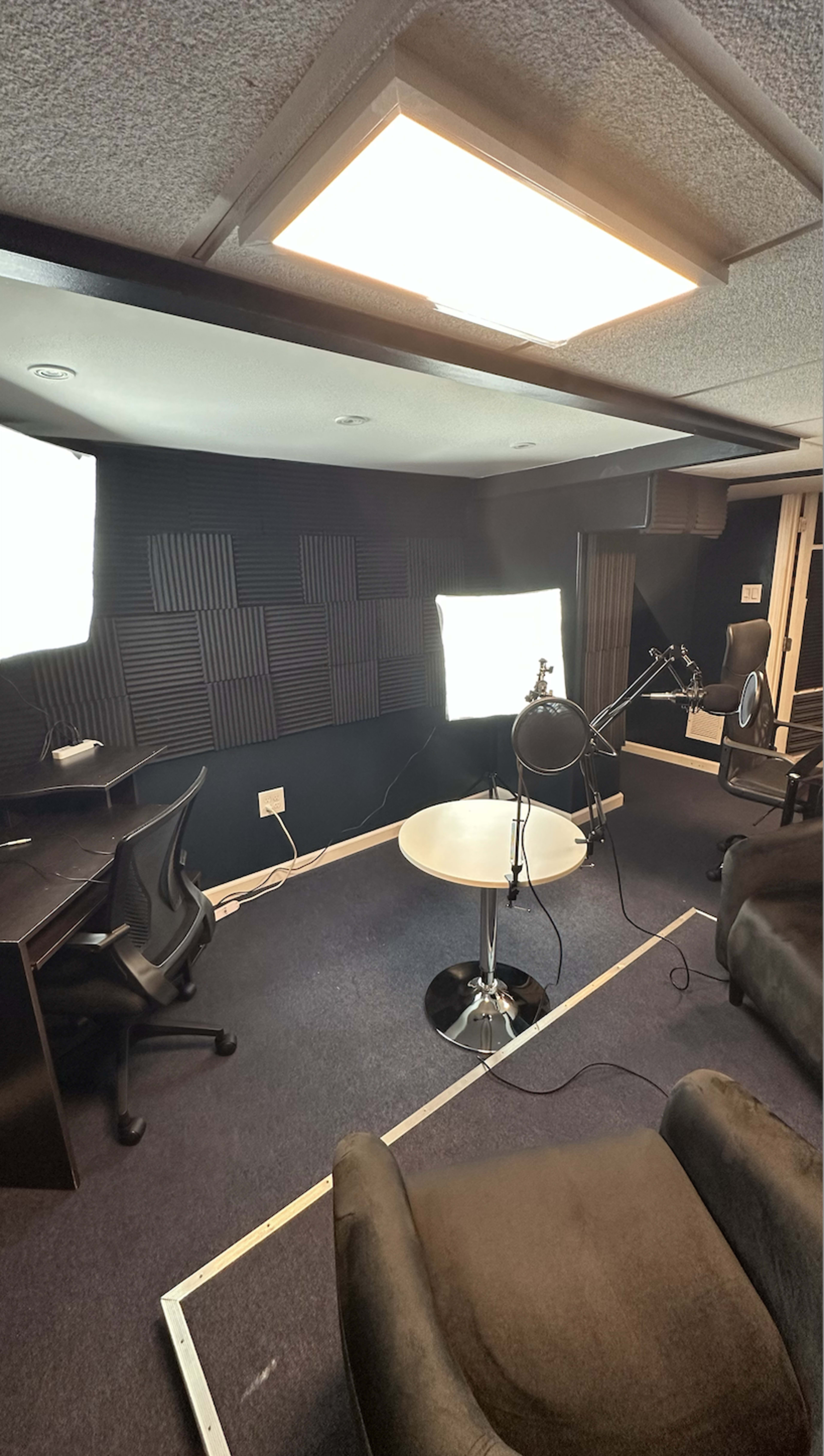 A recording studio with black soundproofing foam on the walls, a circular table, two microphones on stands, and studio lights positioned around.