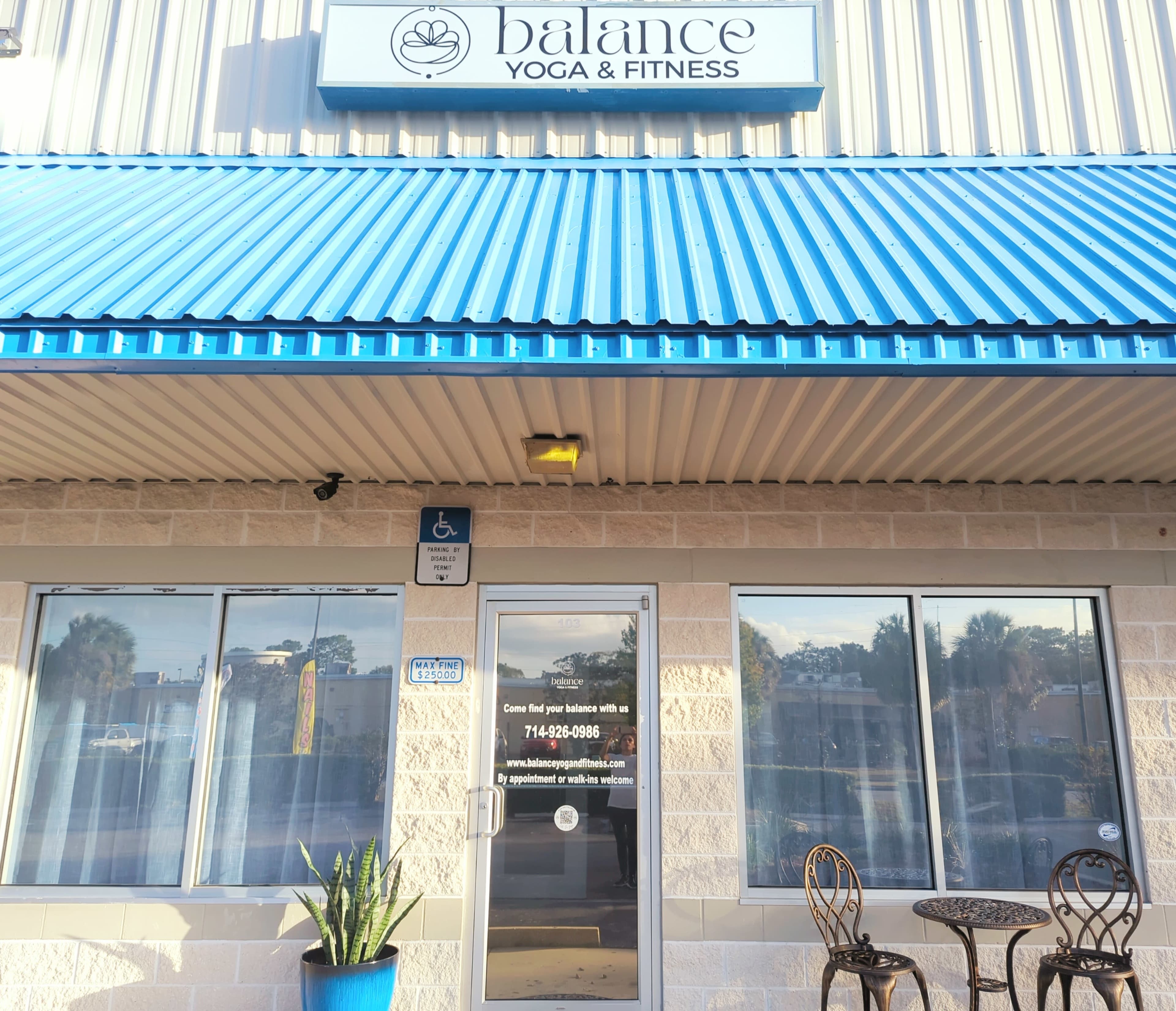 The image shows a fitness studio named "Balance Yoga & Fitness" with large front windows, a blue awning, and a small outdoor seating area.