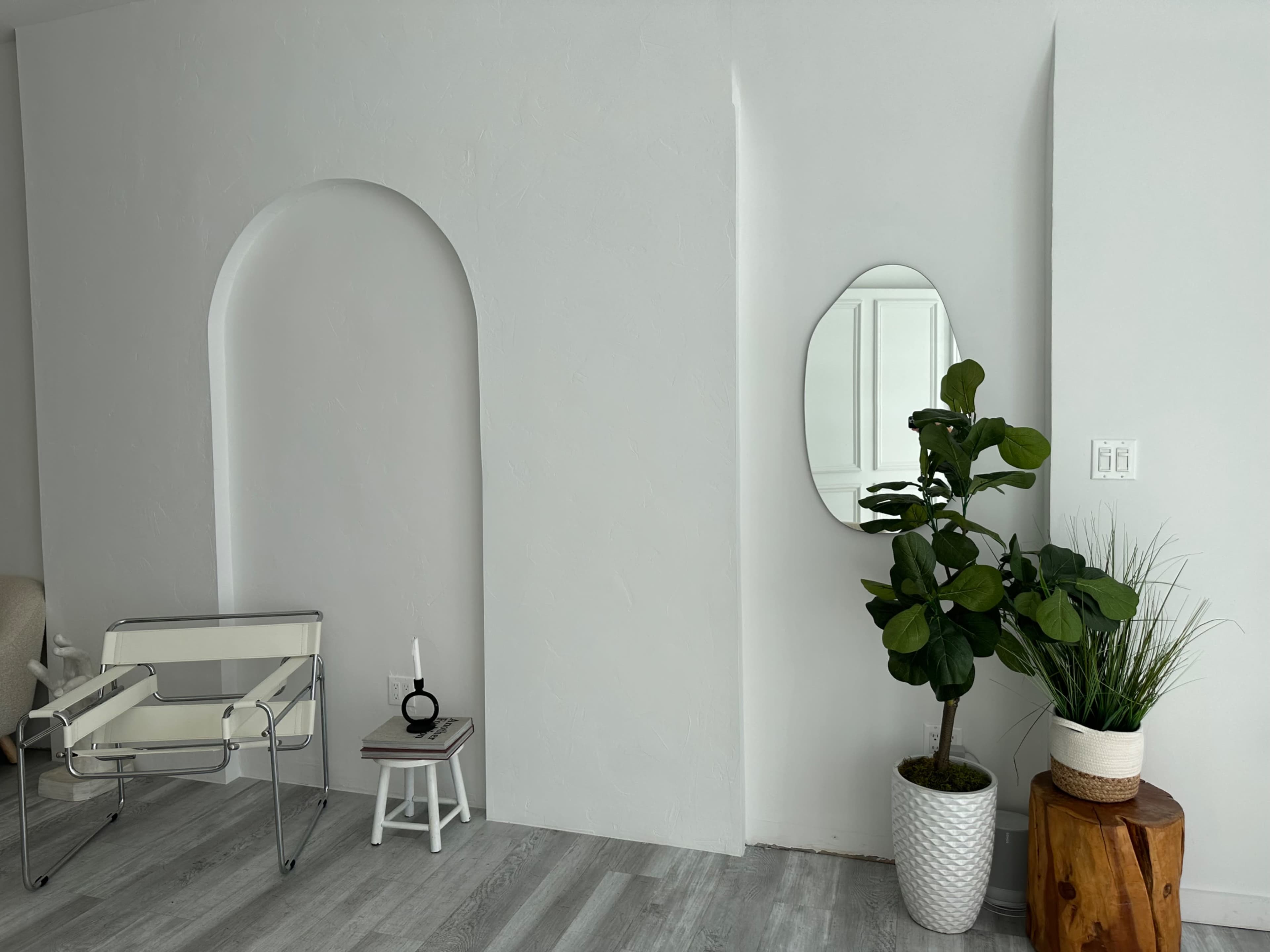 A minimalist interior featuring a white wall with an arch, a mirror, a potted plant, and a small table with a candle.