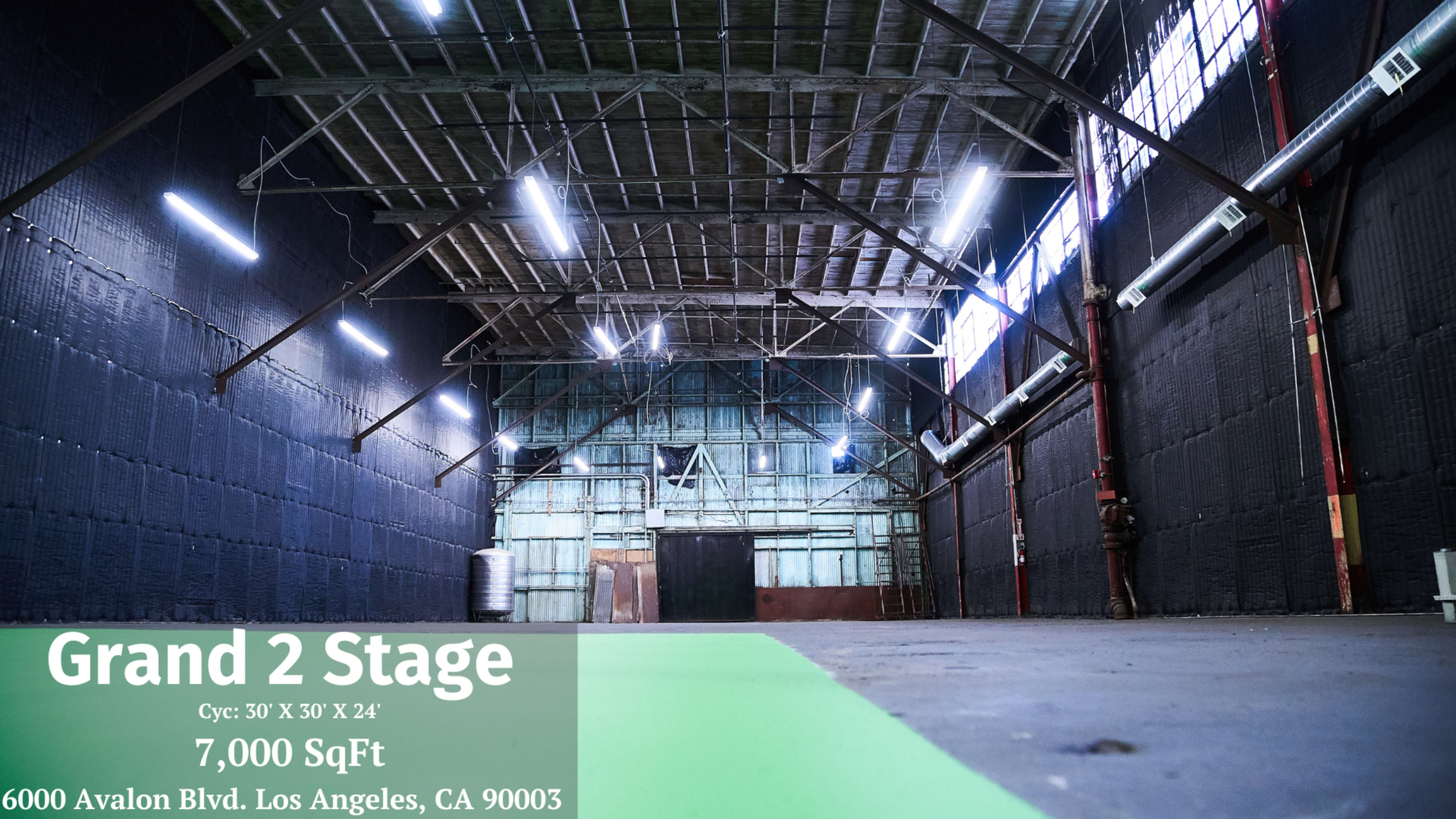 The image shows an empty warehouse space with large windows and exposed beams, marked as "Grand 2 Stage" with dimensions and location details in the corner.