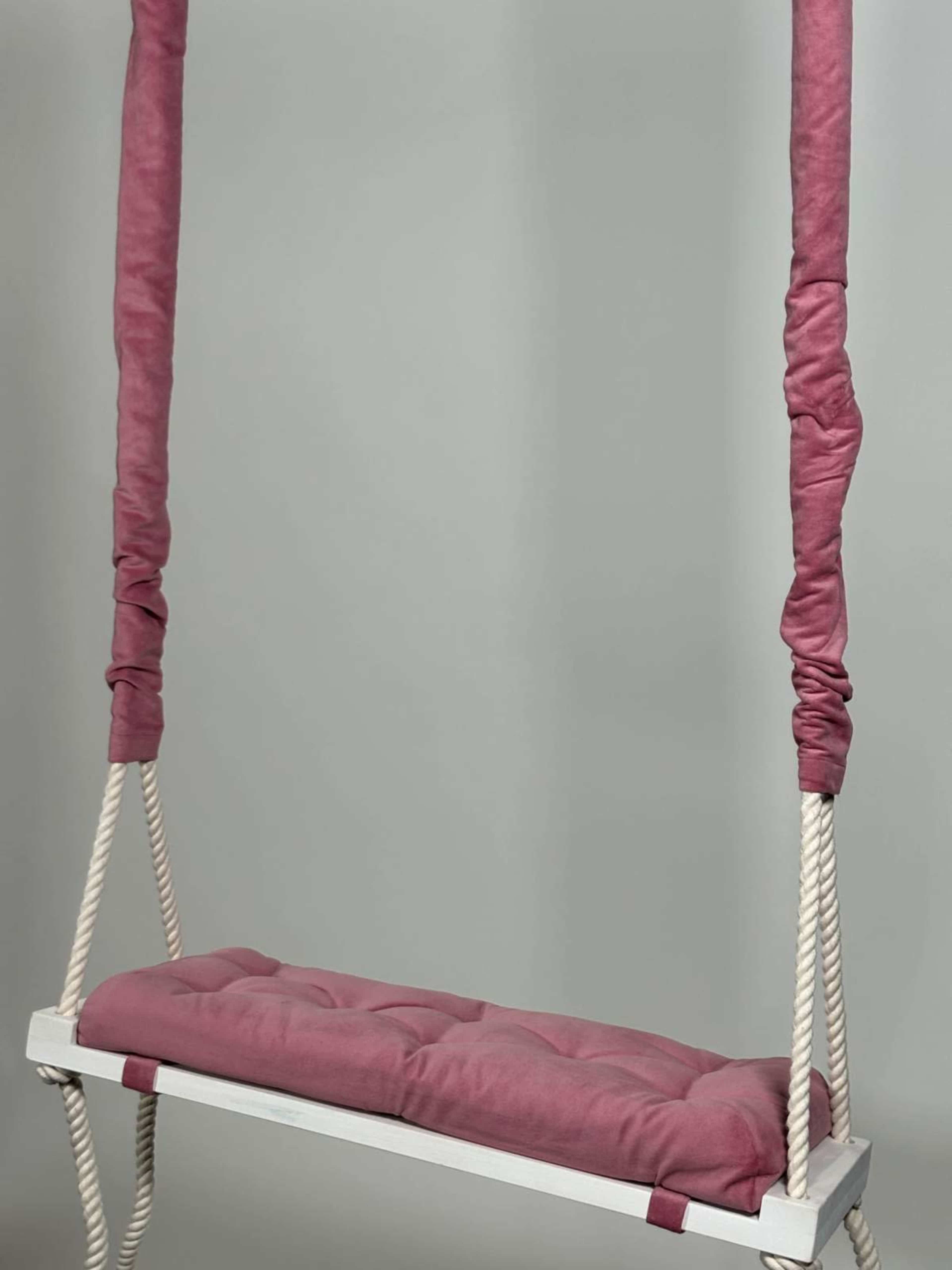 A swing with a tufted cushion in a pink cover is suspended by thick ropes against a gray background.