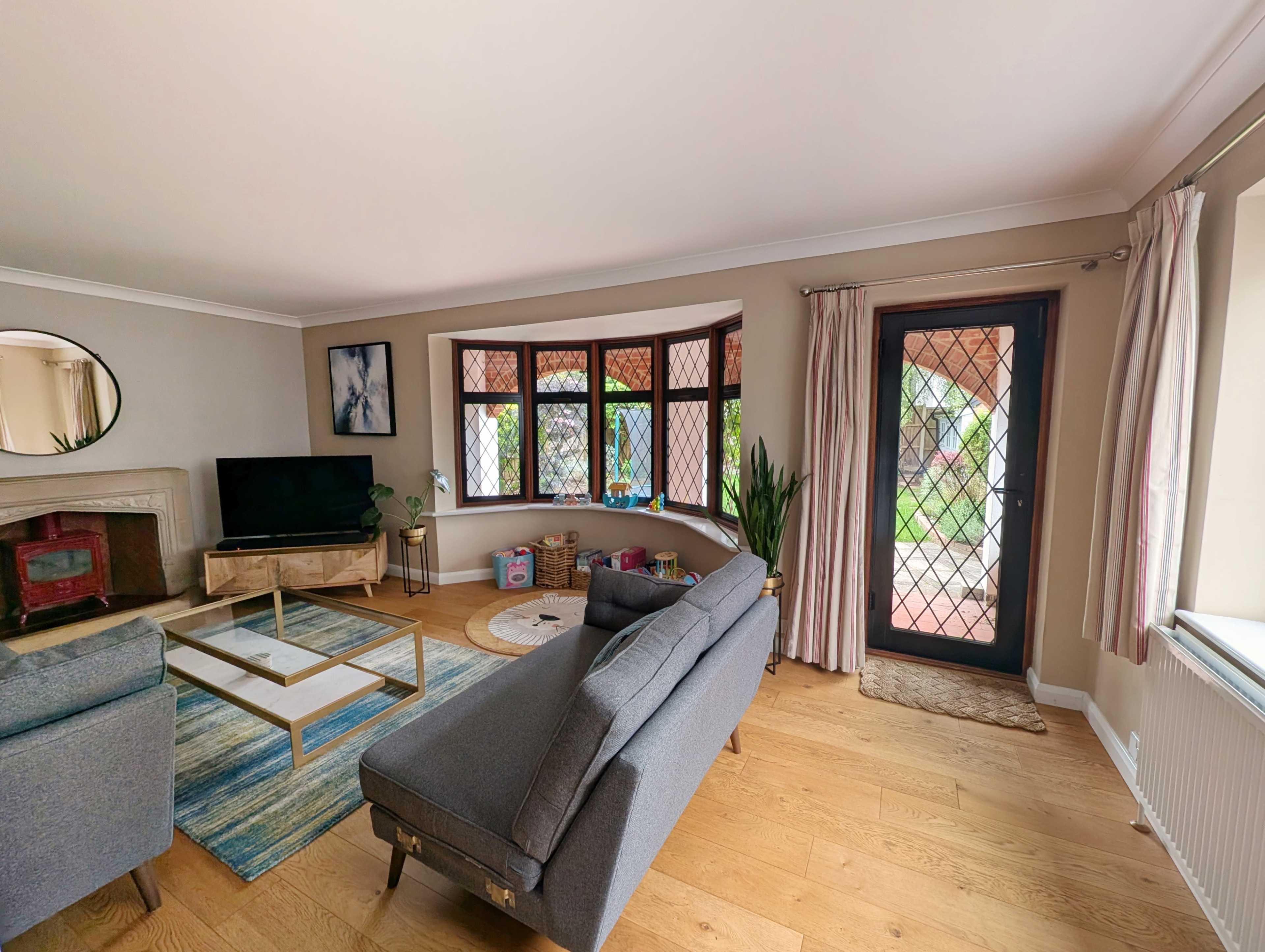The image shows a living room with a gray sofa, a television mounted on the wall, a coffee table in the center, and a door leading to an outdoor area.