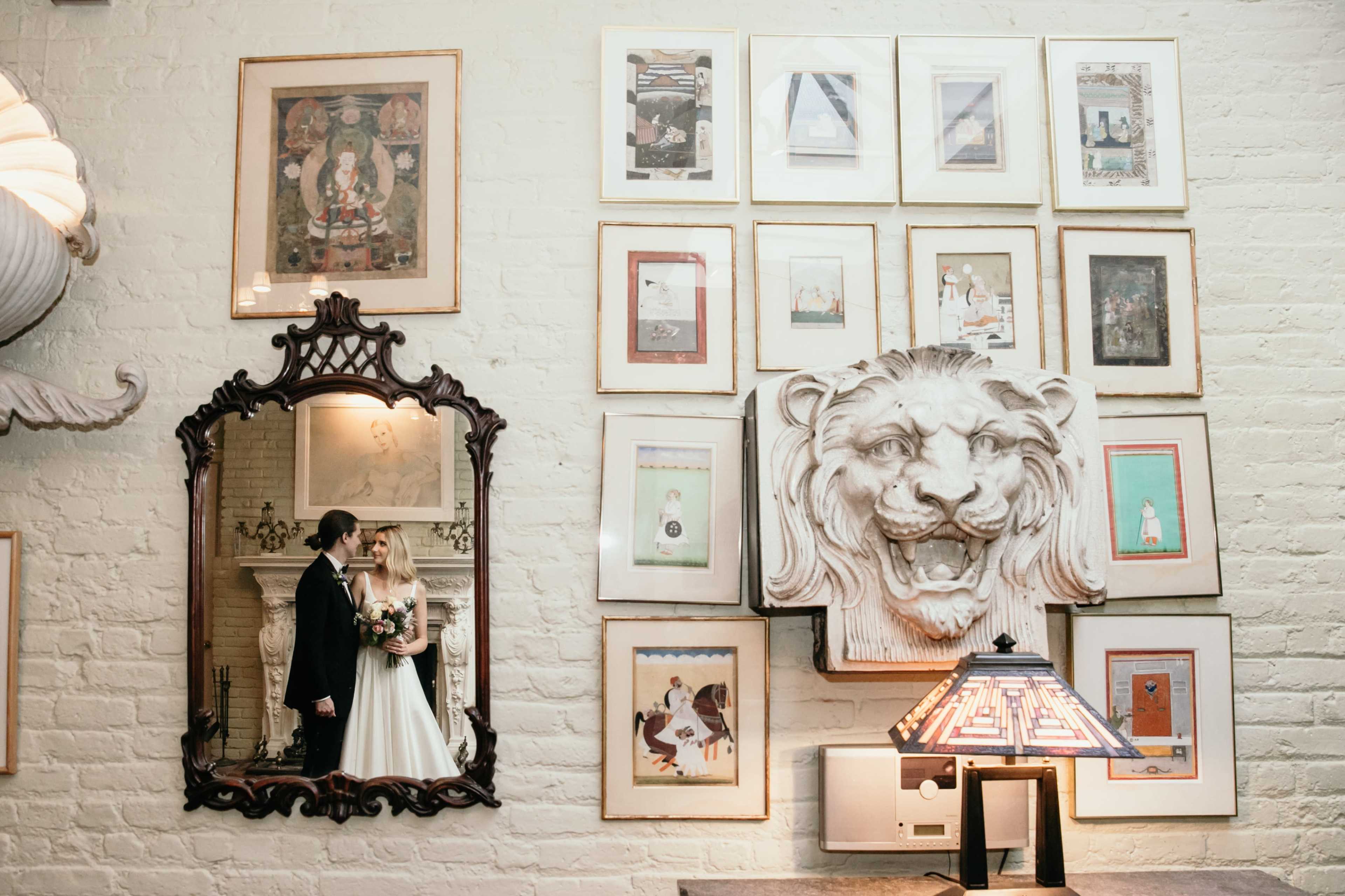 A couple stands in front of a gallery wall adorned with framed artwork and a lion sculpture, reflected in an ornate mirror.