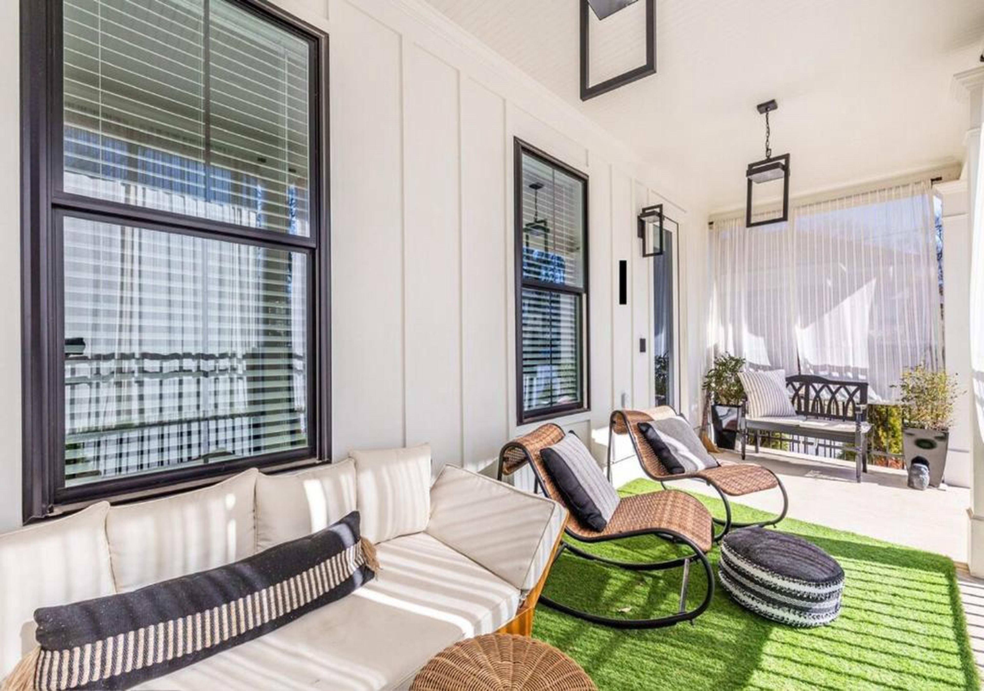 The image shows a covered porch featuring modern seating, a green area rug, and light-filtering curtains.