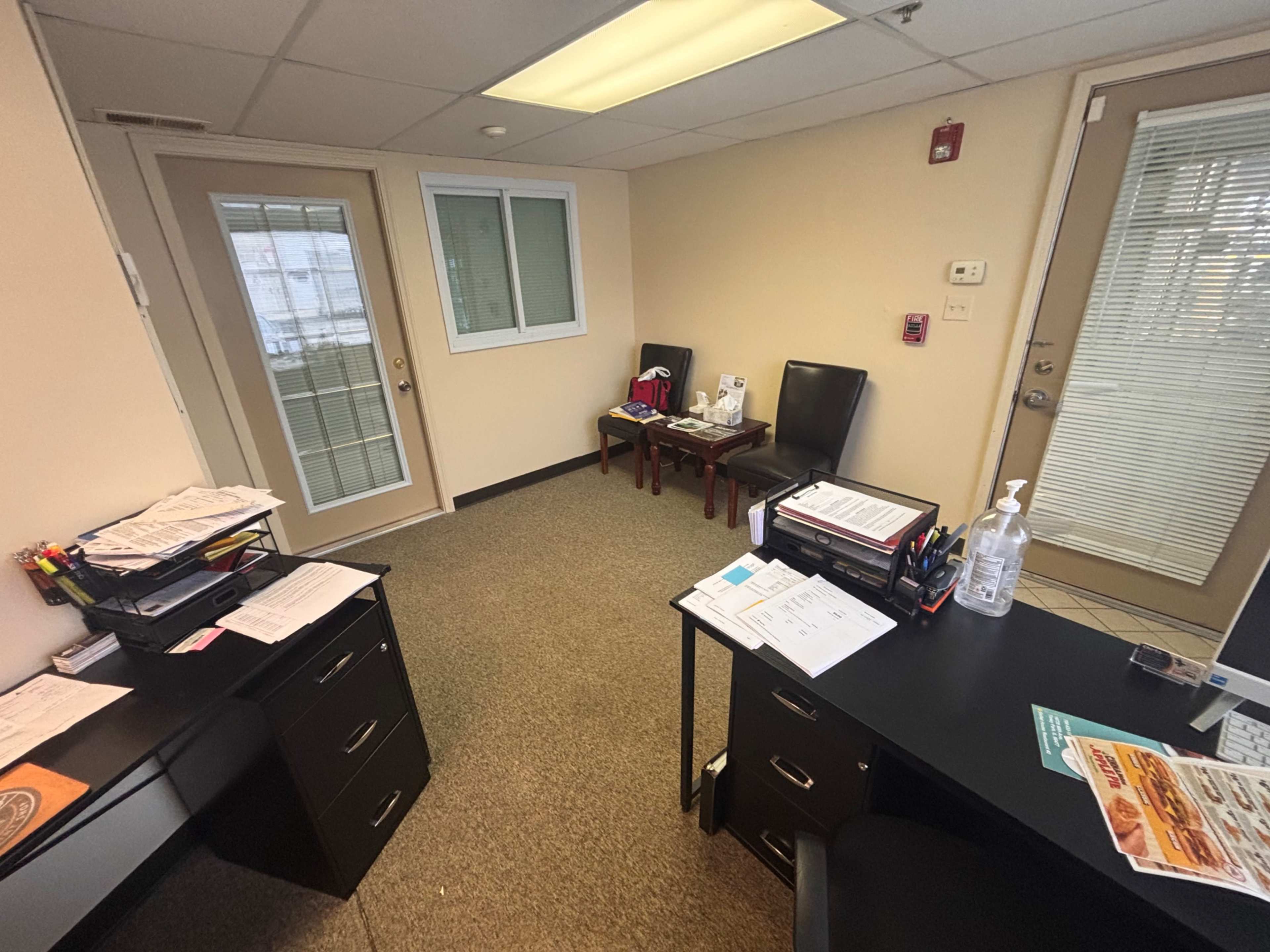 The image shows an office space with two doors, a desk with paperwork, and two empty chairs next to a small table.