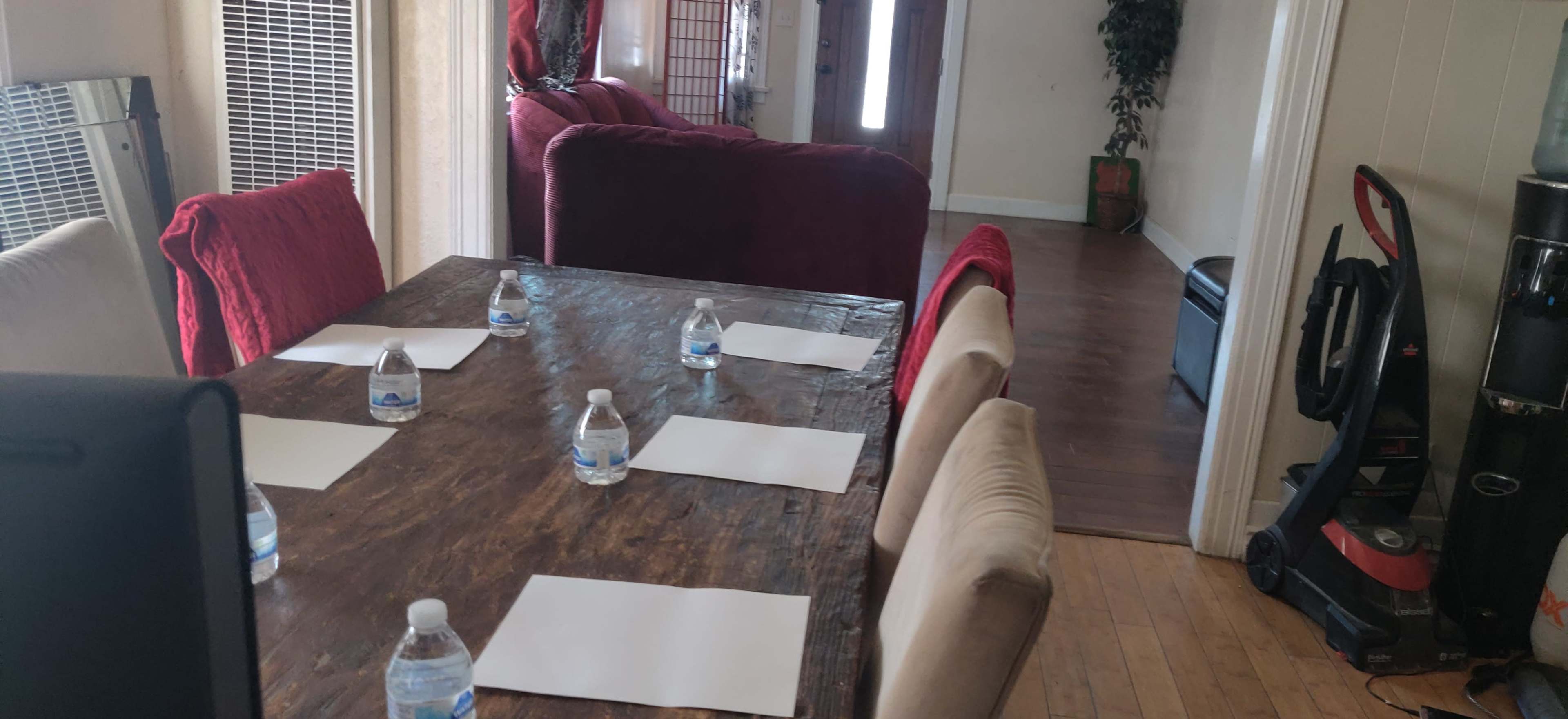 A wooden dining table is set with six paper sheets and water bottles, surrounded by chairs, in a room featuring a couch and a view of an open doorway.