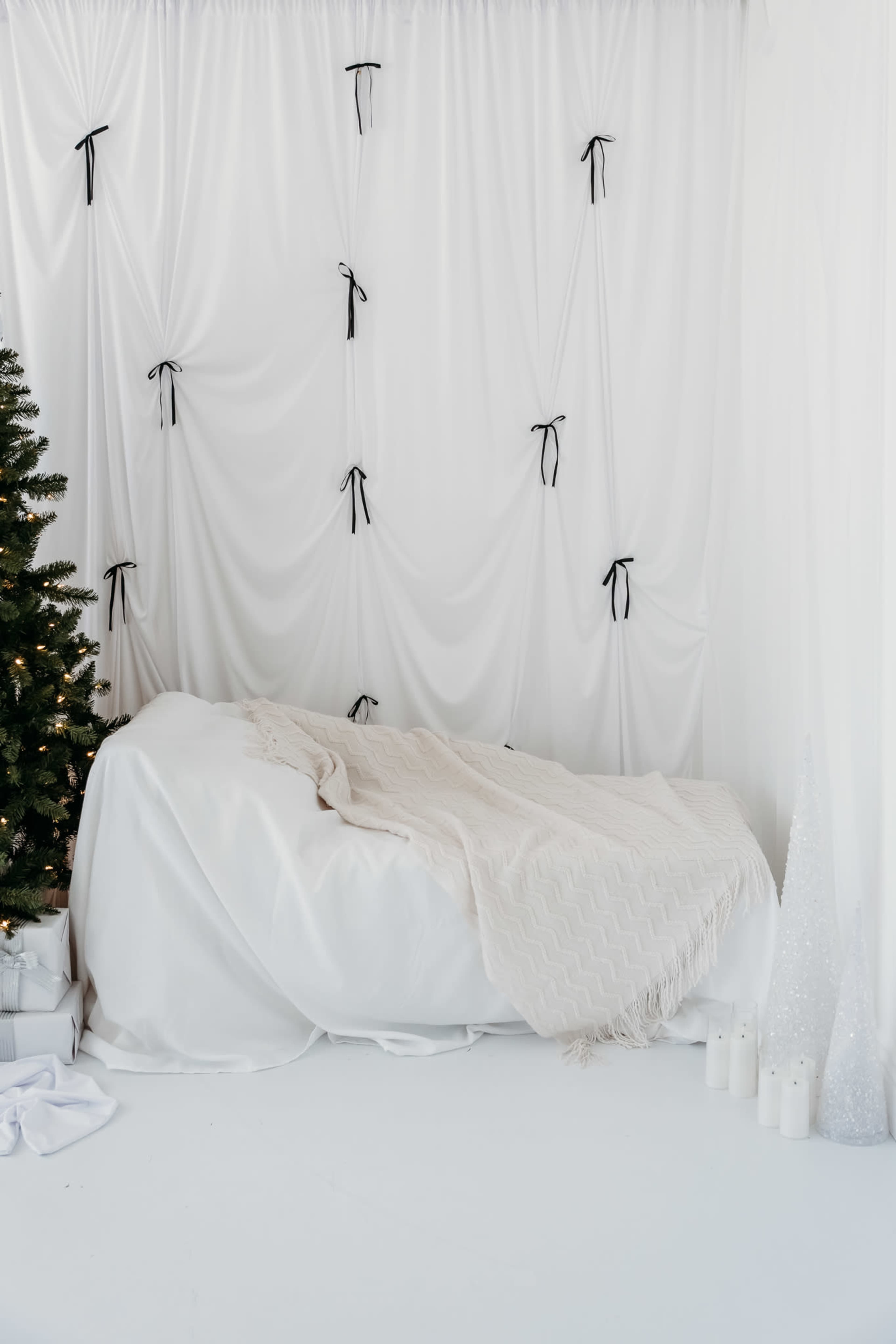 The scene features a minimalist bedroom with a white bed covered in a soft blanket, set against a white backdrop adorned with ribbons, and a Christmas tree nearby.