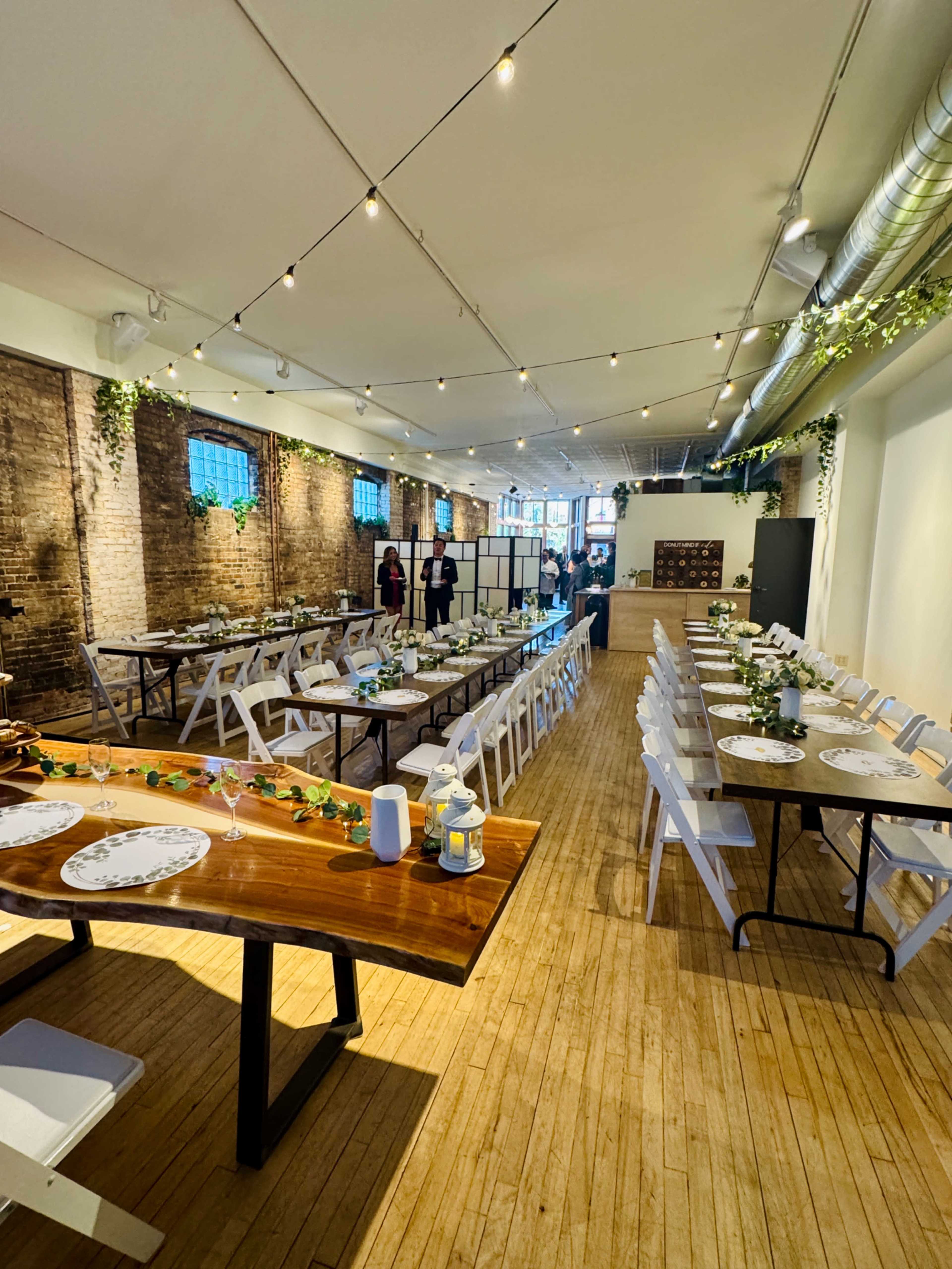 The image shows a spacious event venue set up with long tables for a dining arrangement, decorated with greenery and string lights hanging overhead.