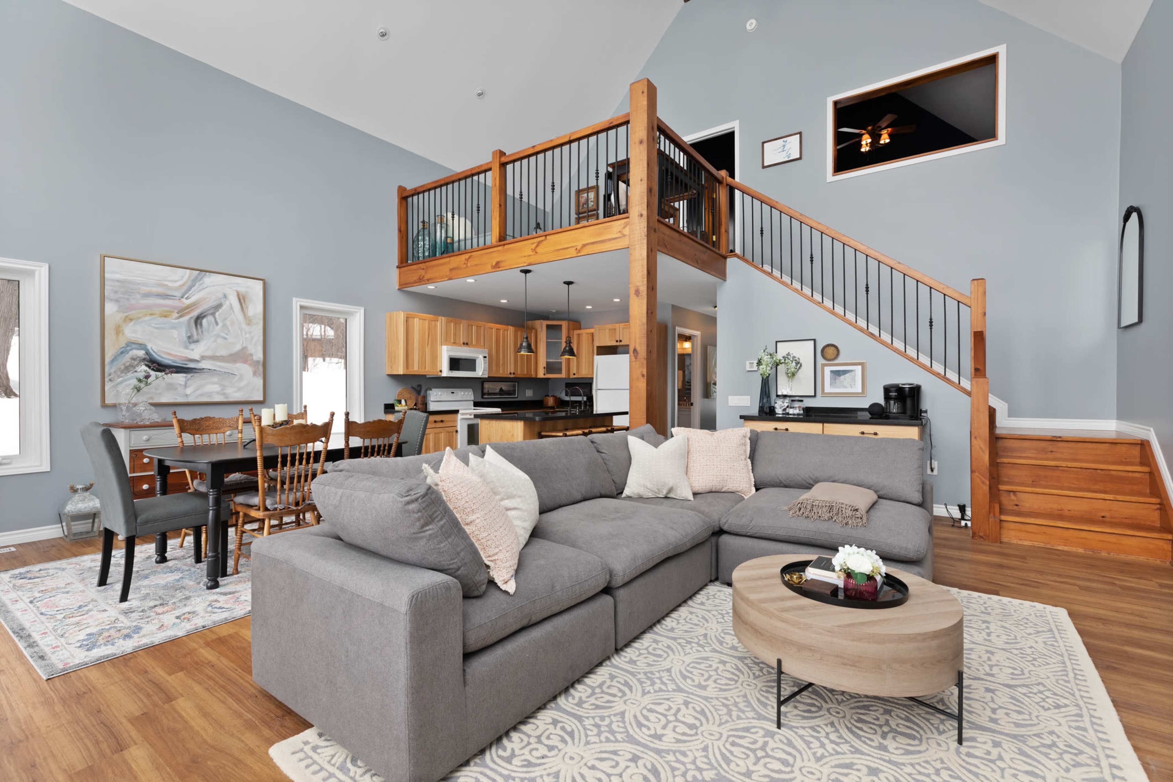 A spacious living area with a gray sectional sofa, a round coffee table, and a dining table adjacent to a staircase leading to a lofted space.