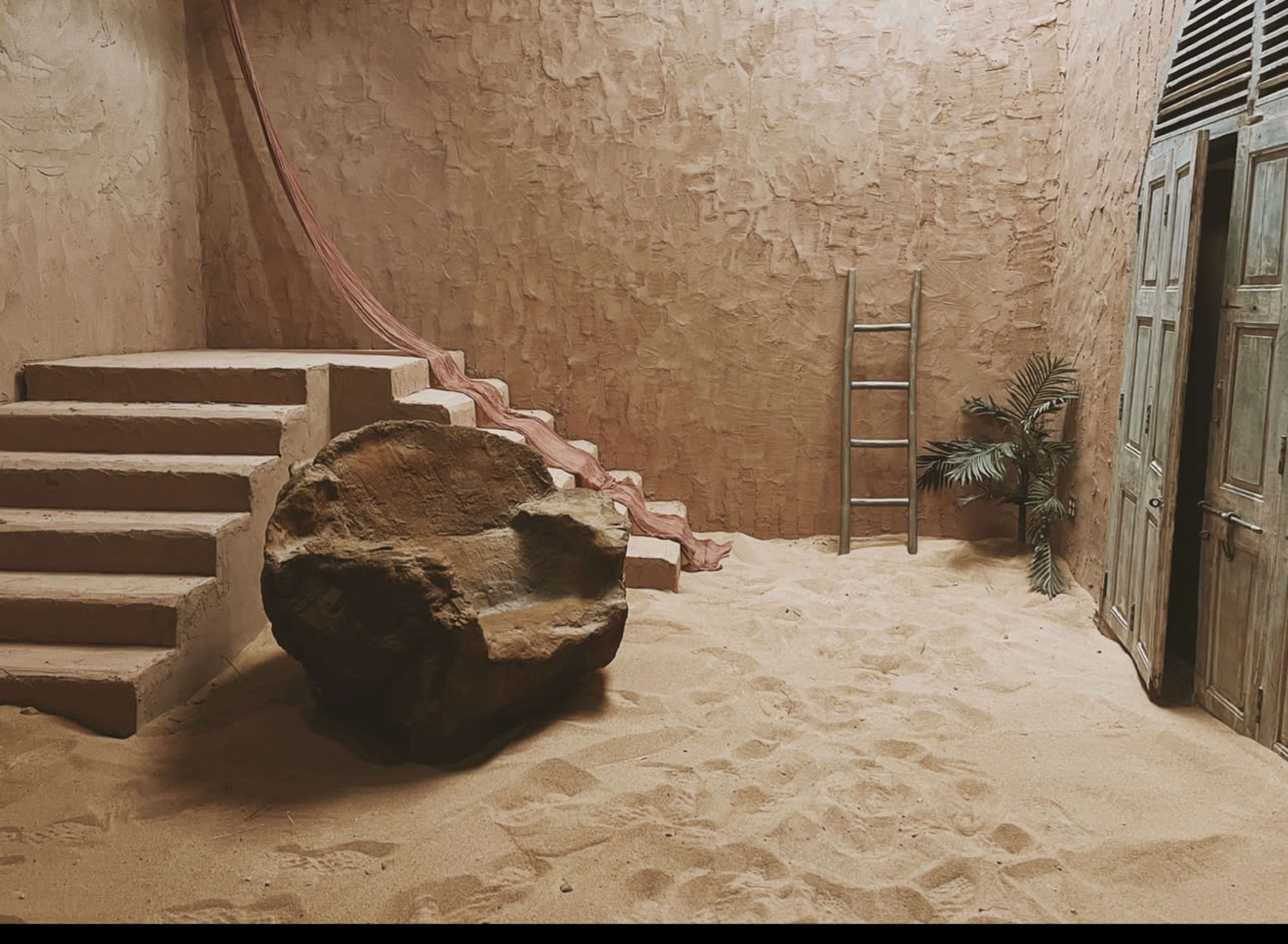 The scene features a sandy interior with a large rock, a ladder against a wall, stairs leading up, and a small palm-like plant in one corner.