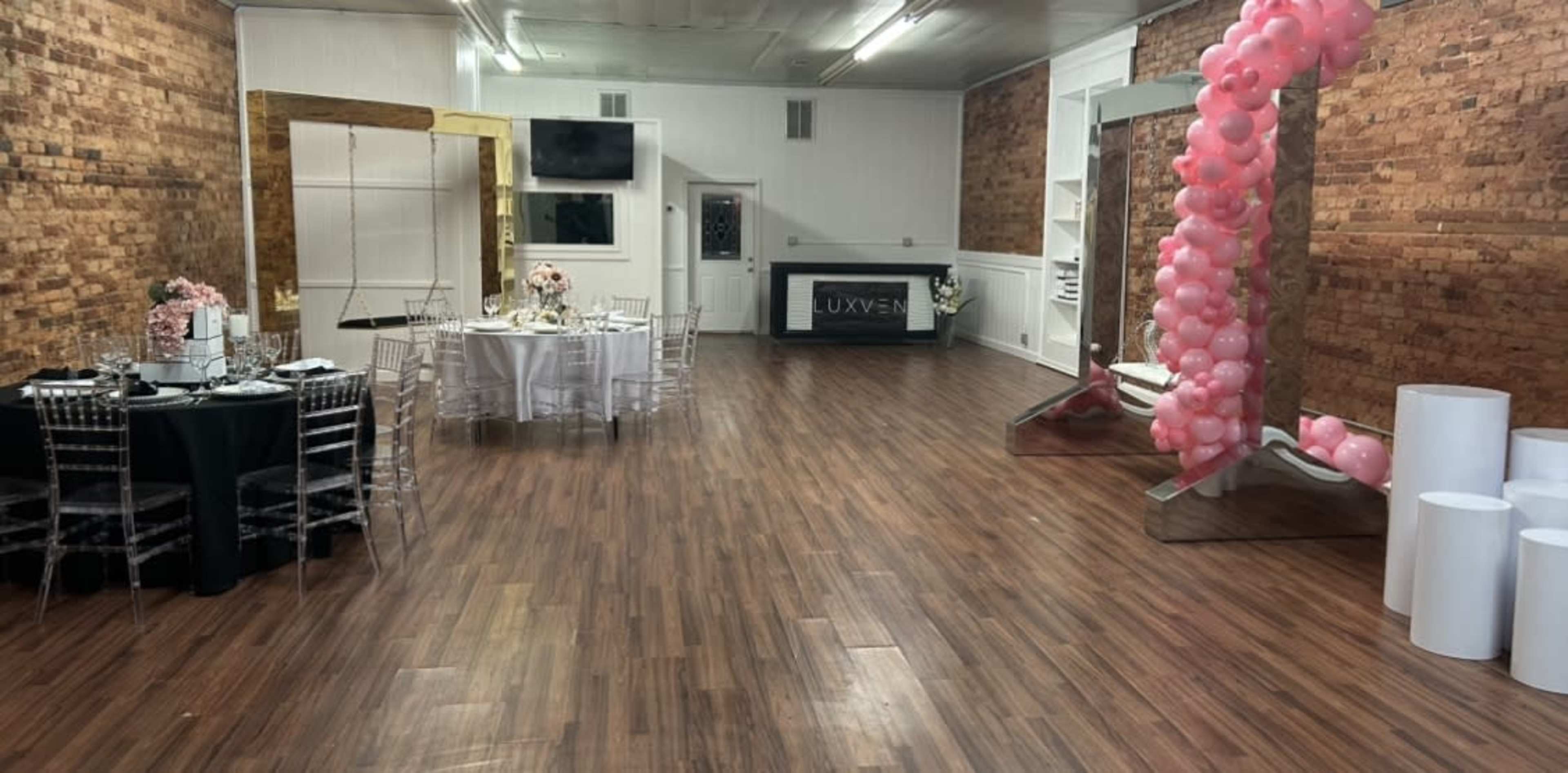 The interior of a venue features wooden flooring, brick walls, two tables set for dining, and decorative elements including a pink balloon arch and mirrored accents.