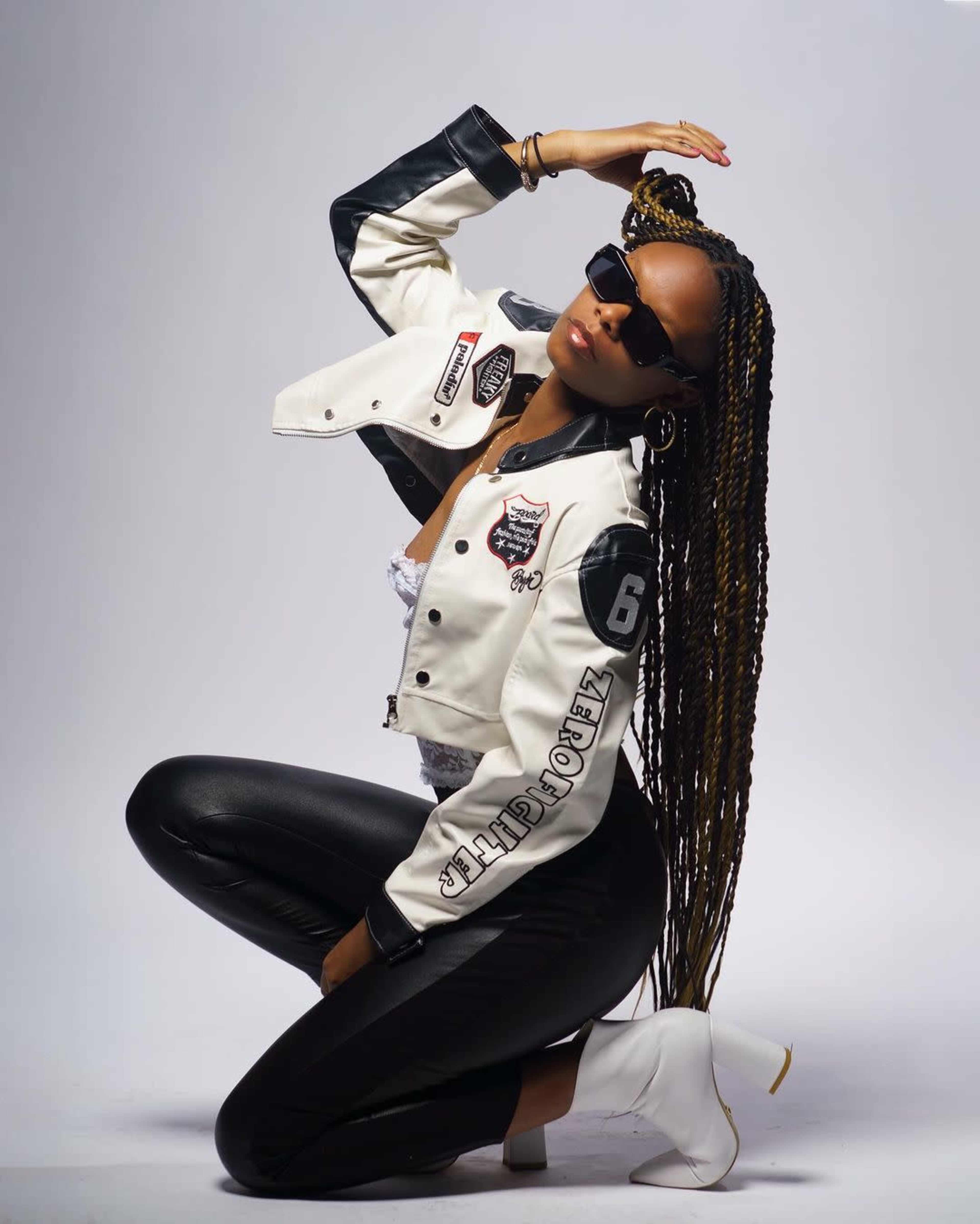 A person poses in a white jacket with black accents and fitted black leggings, wearing sunglasses and stylish ankle boots.