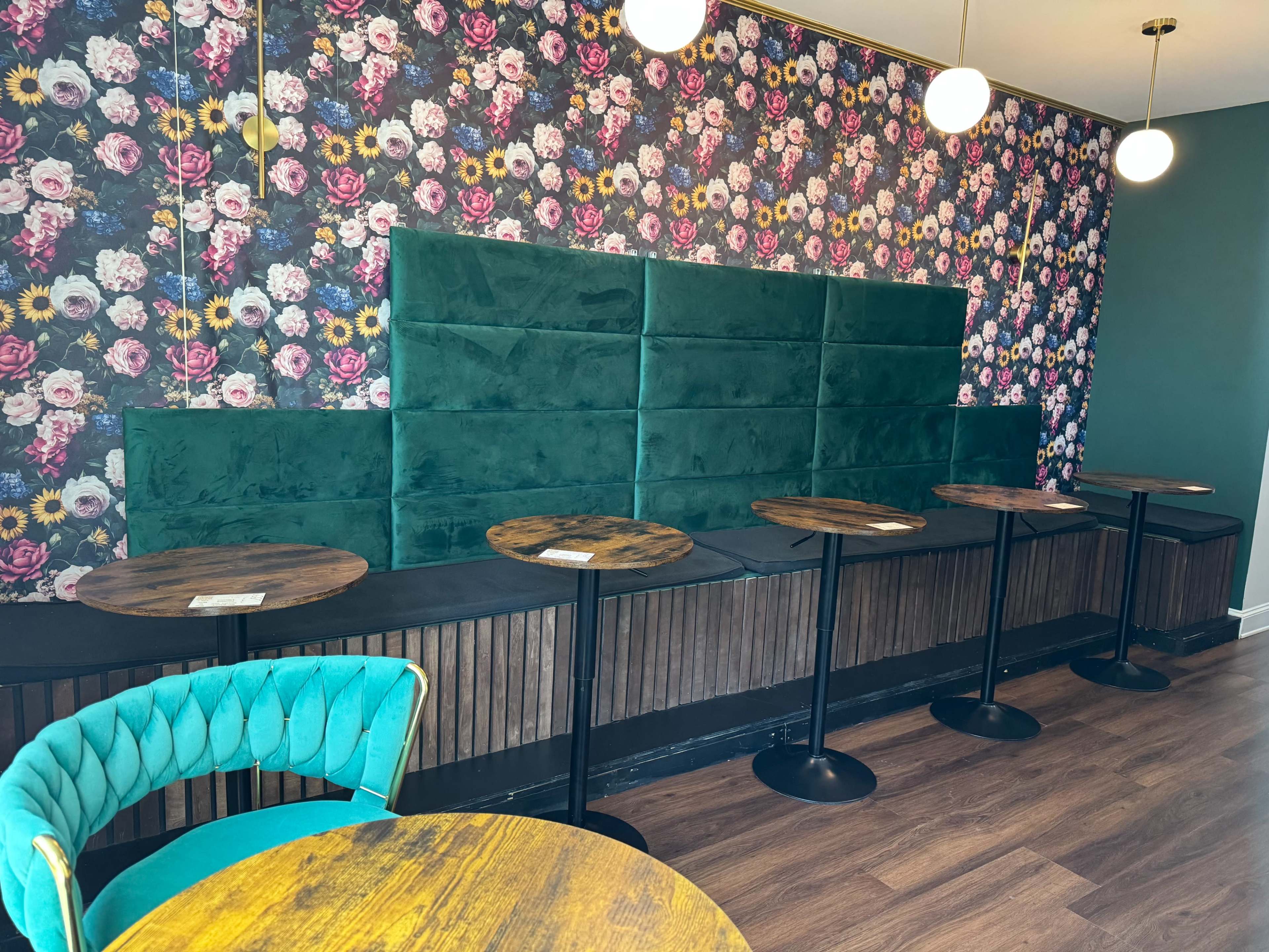 The image shows a cozy café interior featuring a floral wallpaper backdrop, green upholstered seating, and small round tables with black bases.