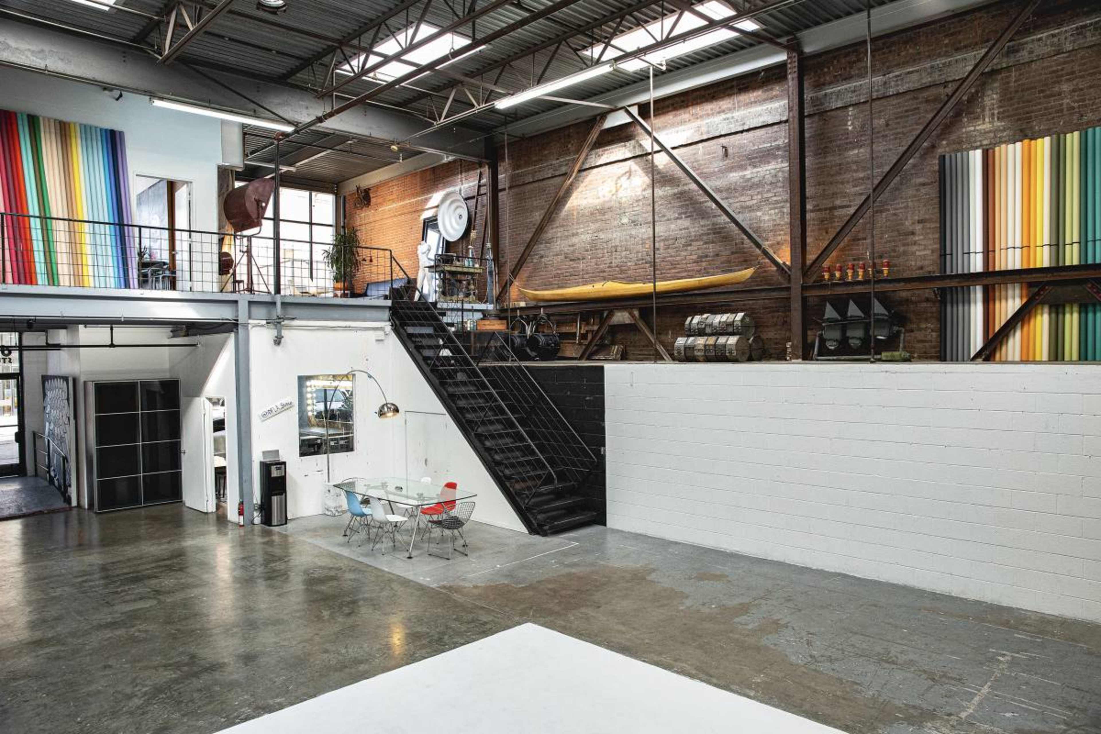 An industrial-style loft with a staircase leading to an upper level, featuring large windows, exposed brick walls, and colorful striped artwork.