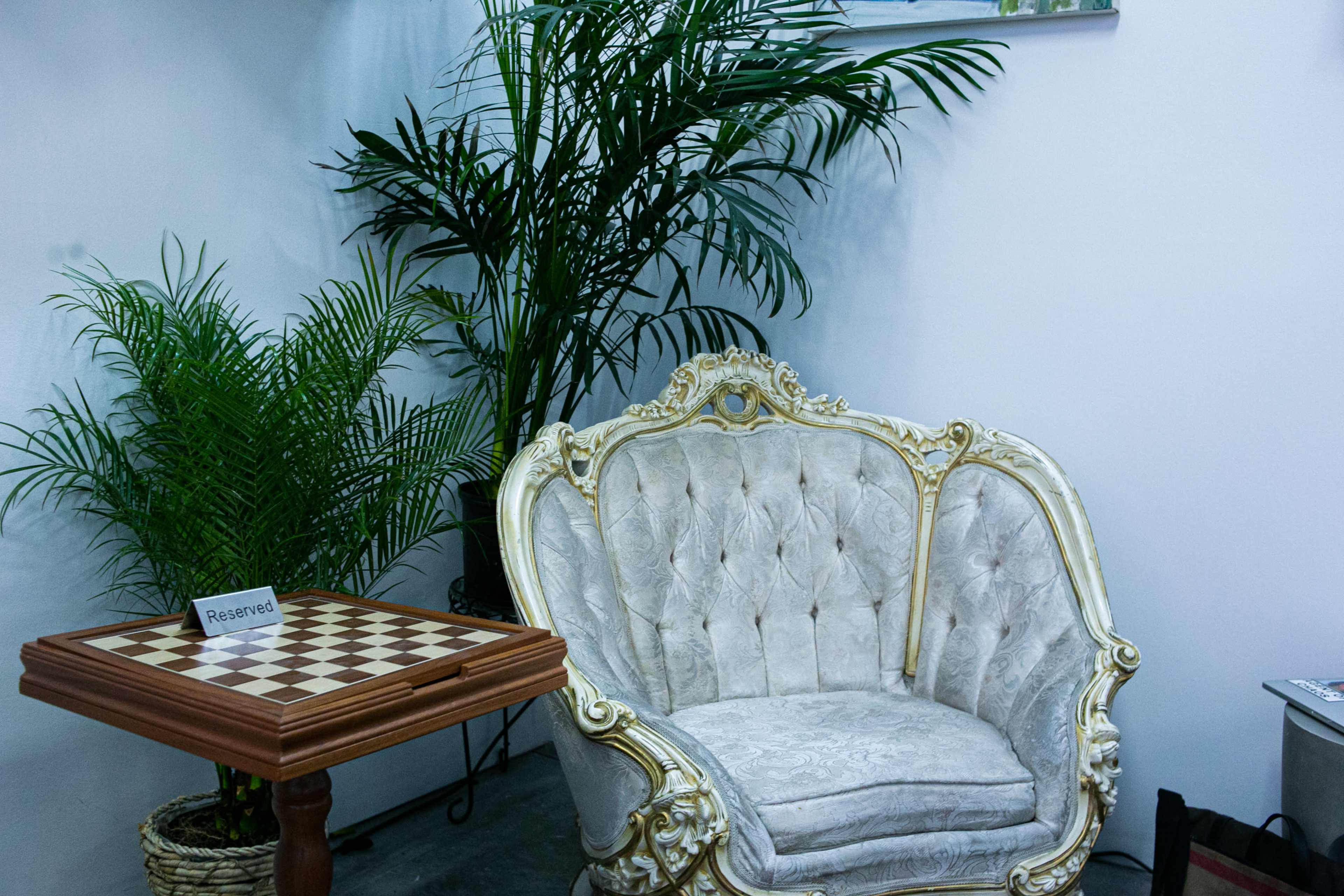 A vintage, tufted armchair is positioned beside a chess table with a "Reserved" sign, surrounded by potted palm plants.