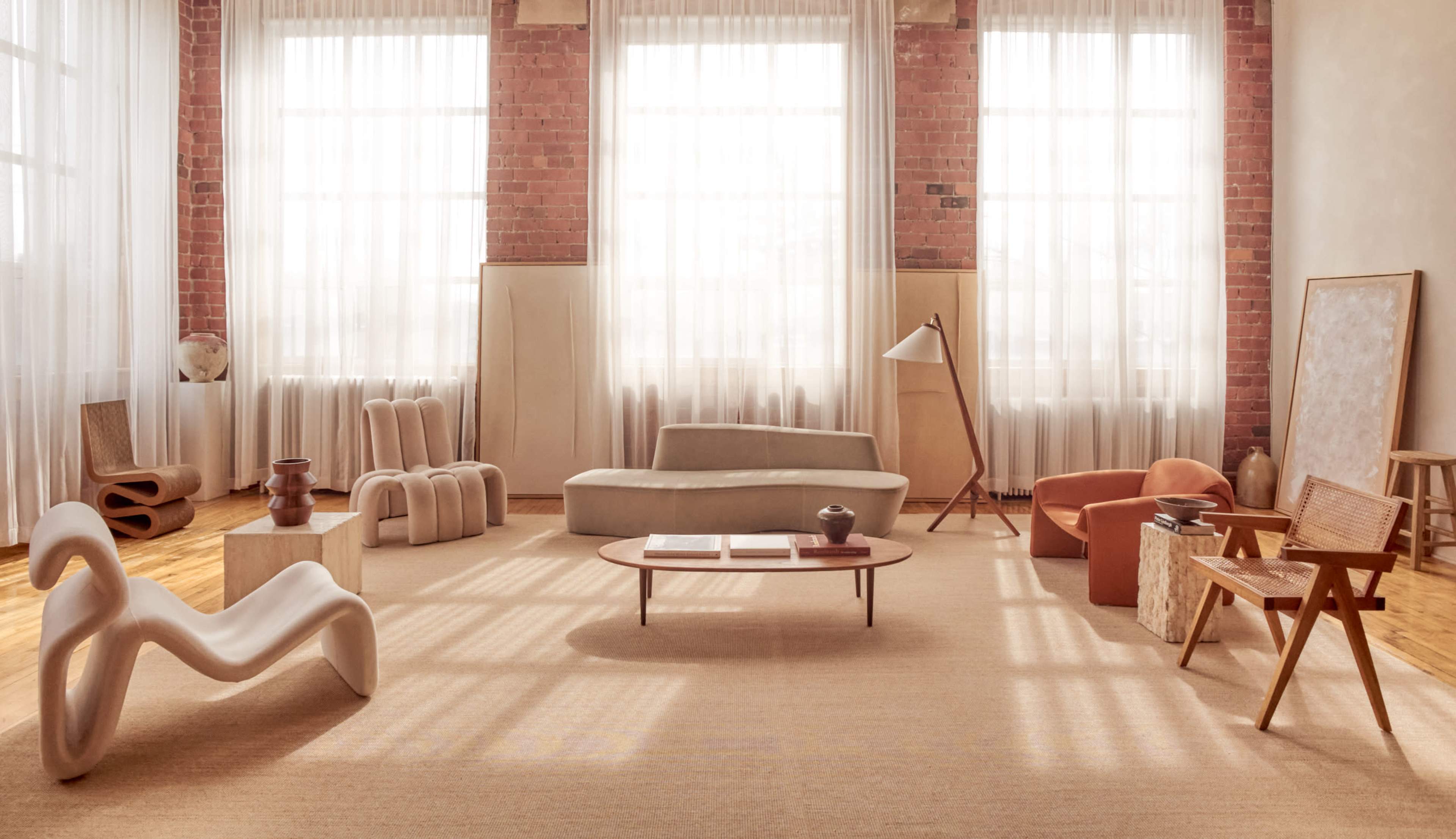The image features a spacious, well-lit interior with large windows, showcasing a modern living area filled with a variety of unique furniture pieces, including a curved sofa, armchairs, and decorative accessories.