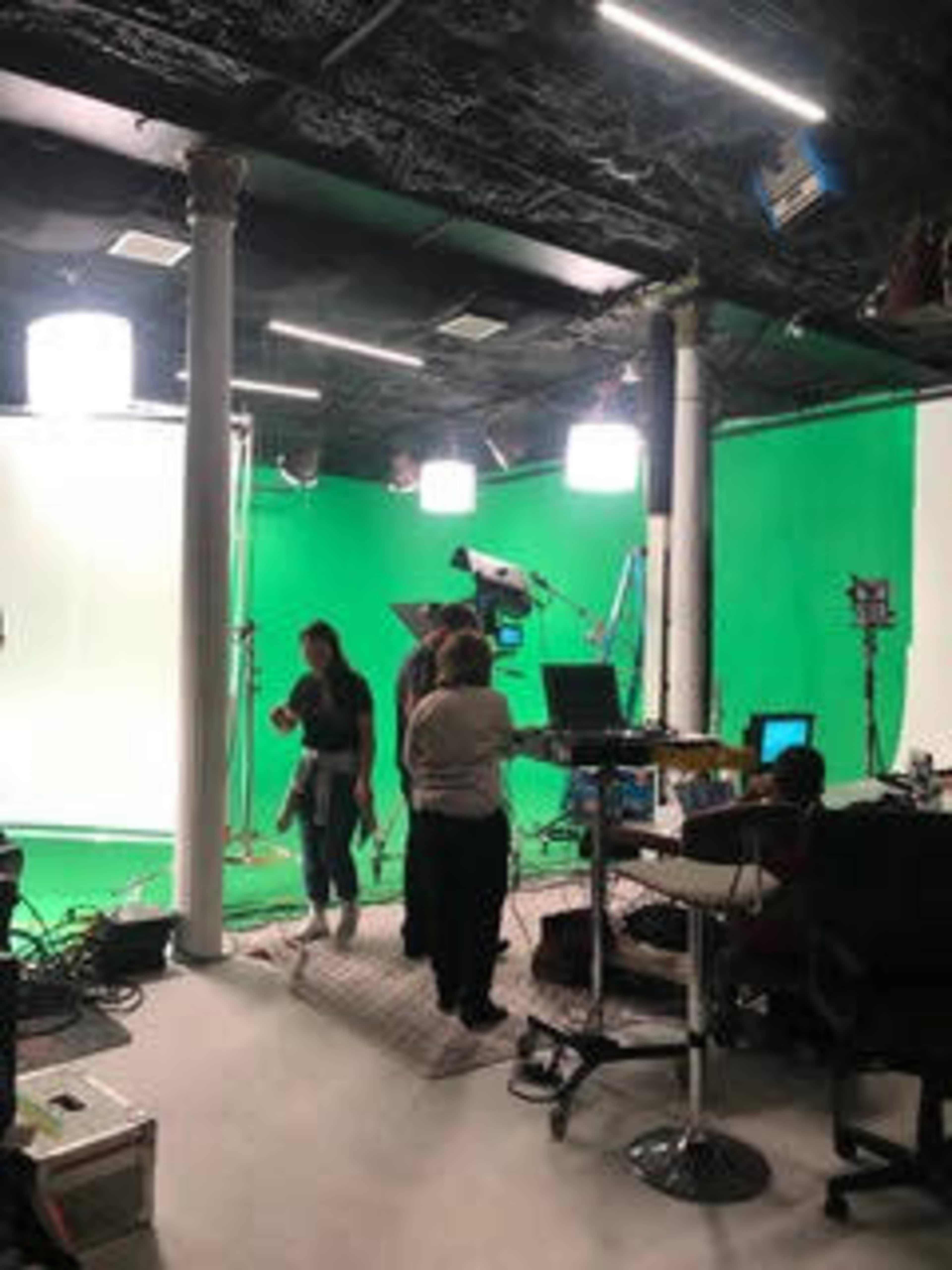 The image shows a filming studio with a green screen setup, bright lighting, and several people working on various tasks.