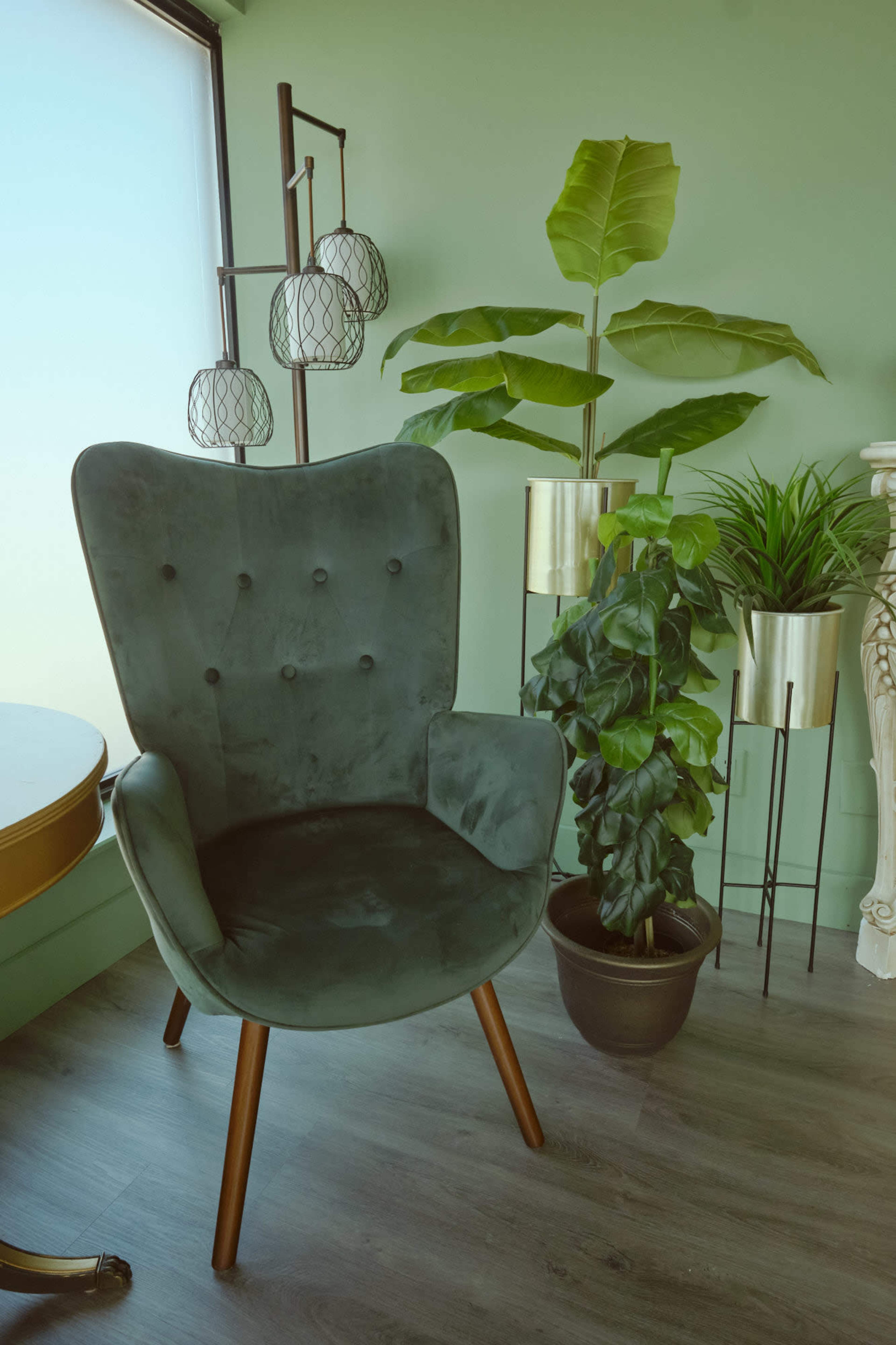A green upholstered chair with wooden legs is positioned next to plants in decorative pots against a light green wall.