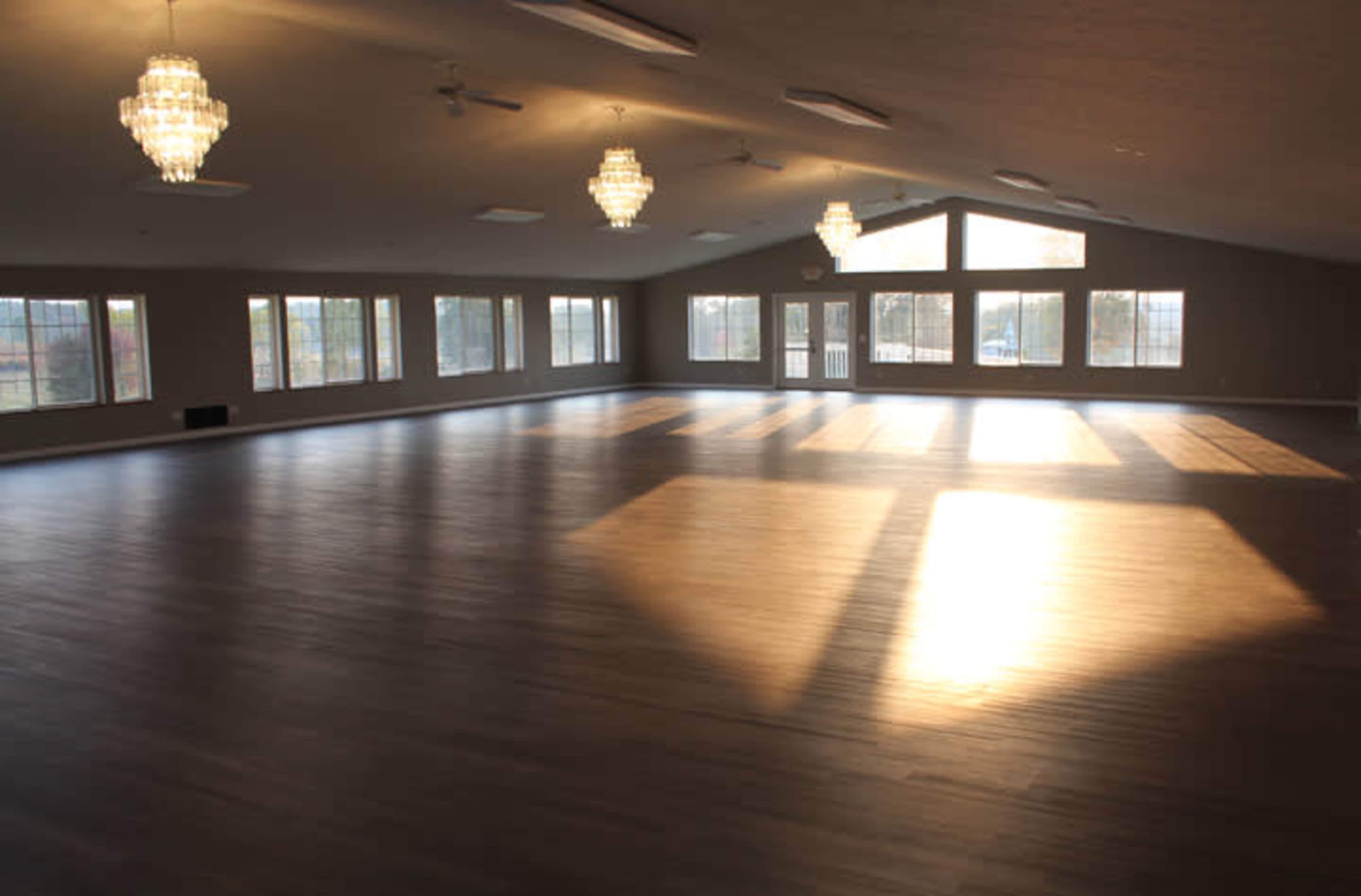 A spacious interior room with large windows allowing natural light to flood in, illuminating the wooden floor and featuring two chandelier fixtures.