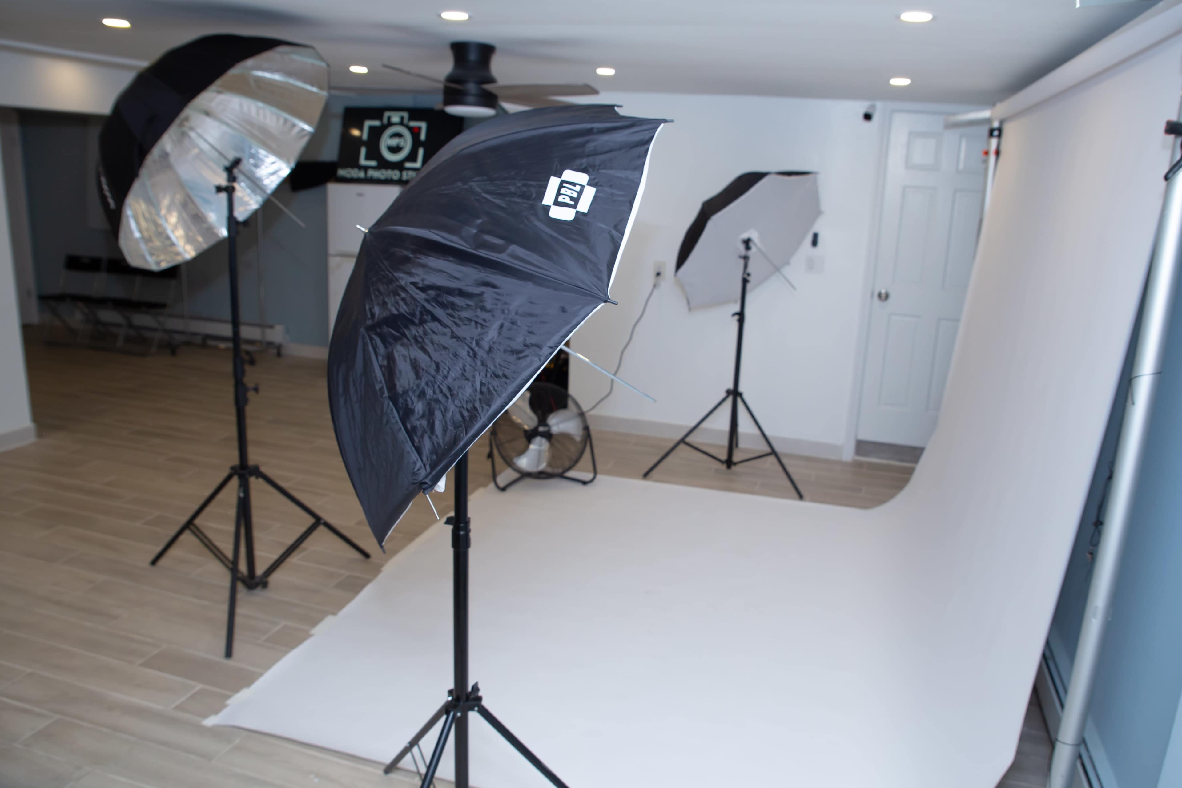 The image shows a photography studio set up with several light umbrellas, tripods, and a white backdrop on the floor.