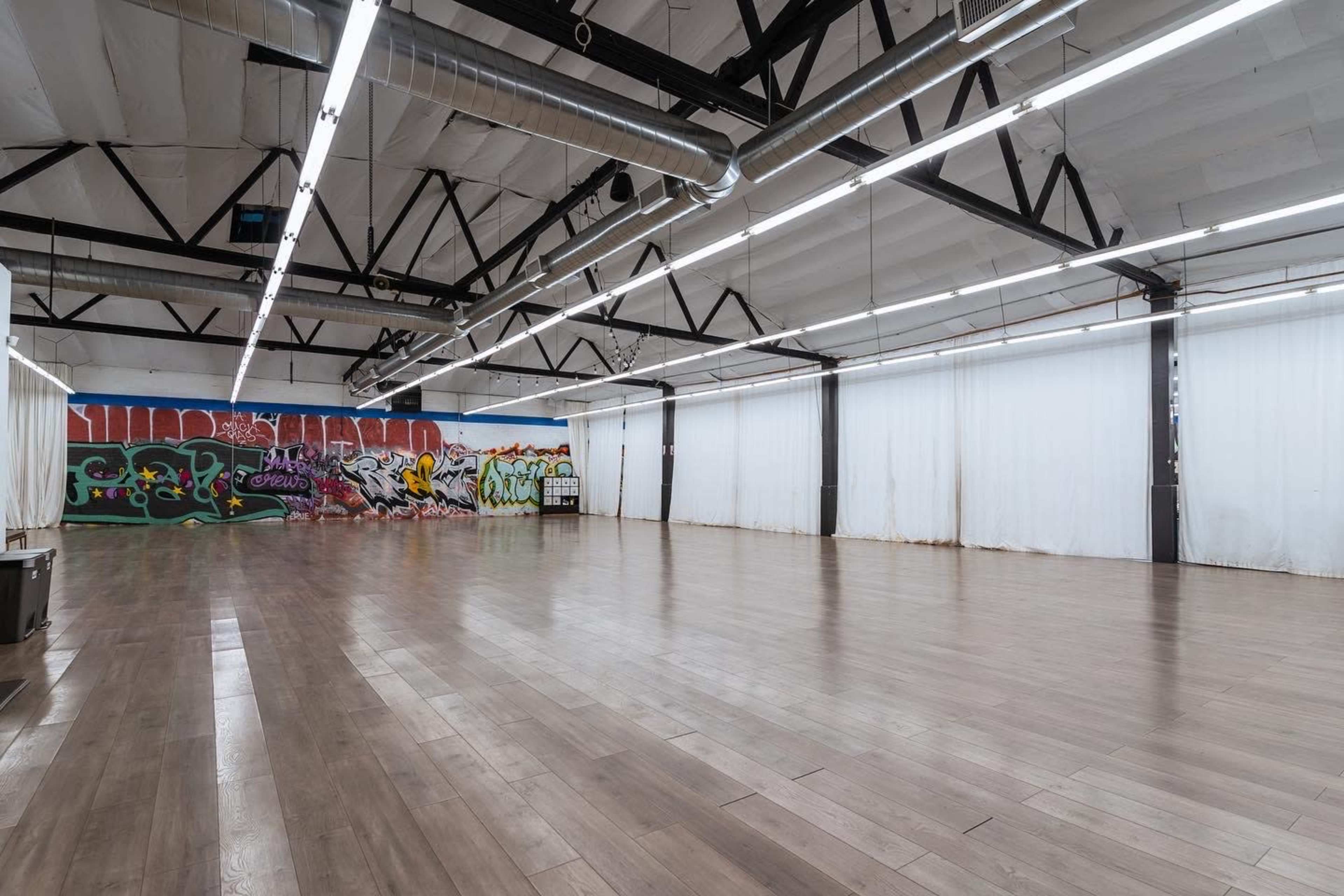 The image shows a spacious room with hardwood floors, high ceilings, and colorful graffiti on one wall, while the other walls are covered with white fabric curtains.