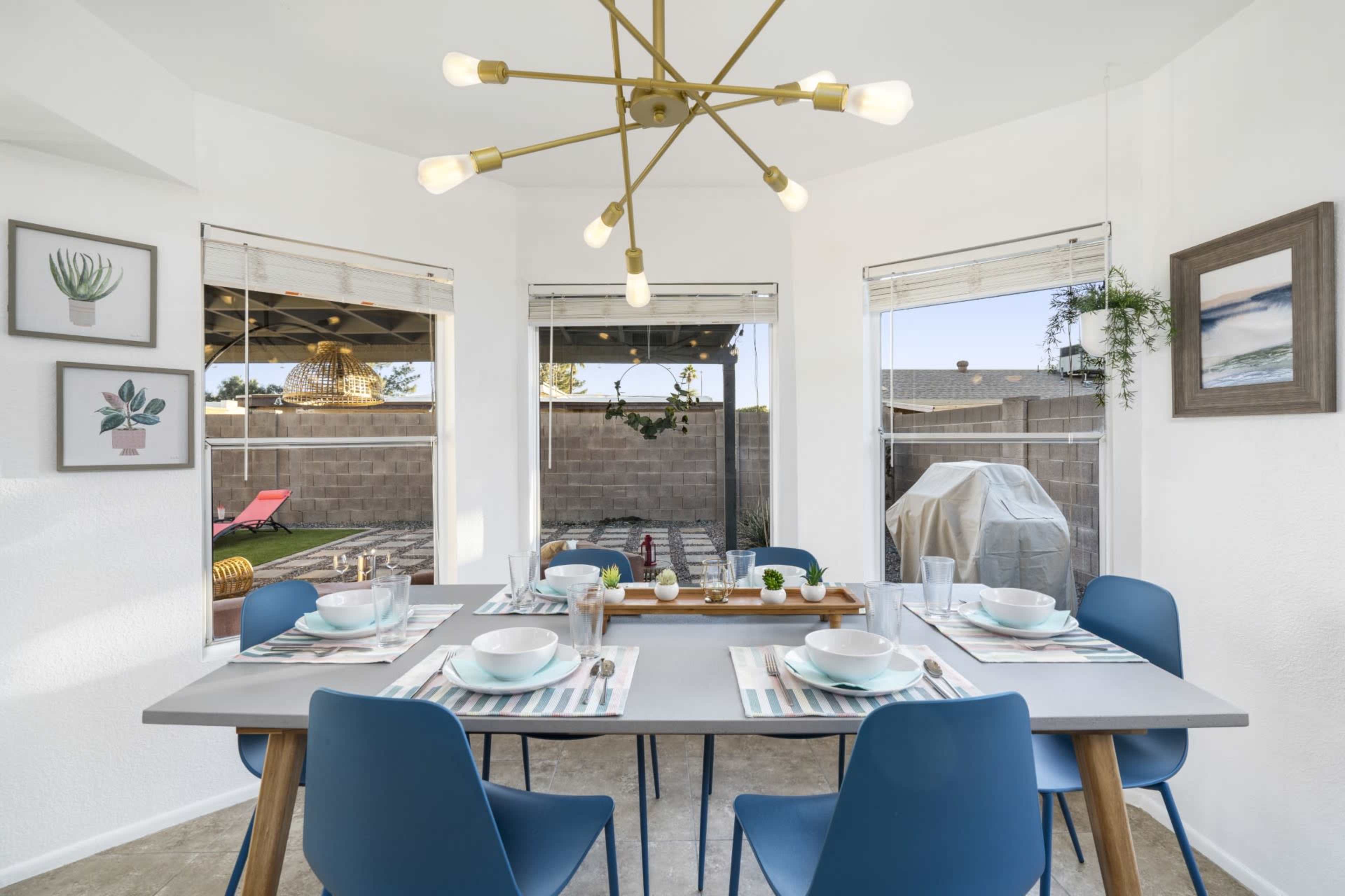 A modern dining area features a table set for six, brightened by a unique light fixture and surrounded by large windows that overlook a patio.