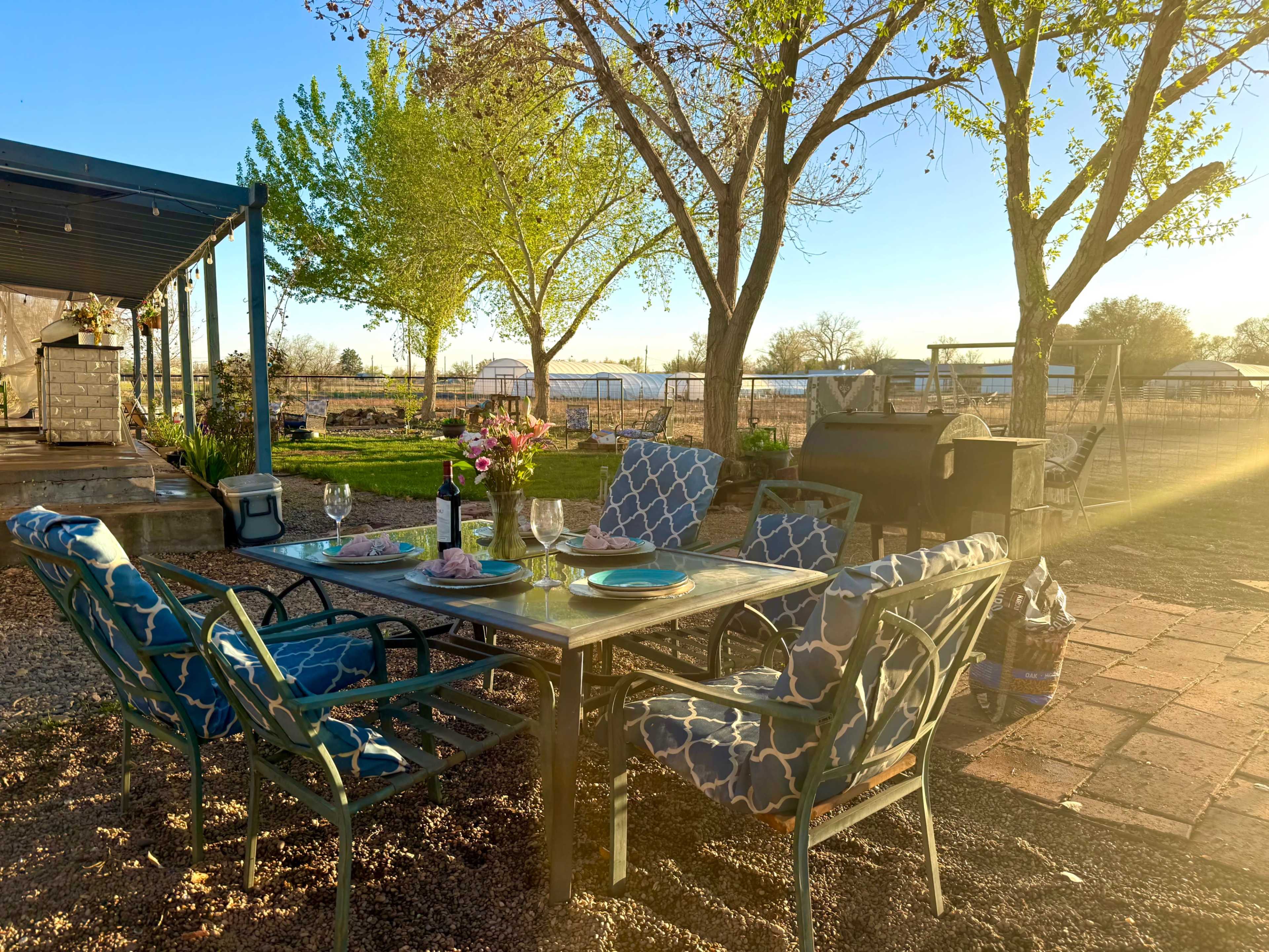 A patio dining area is set with a table and chairs, surrounded by trees and a grass lawn, under a clear sky during sunset.