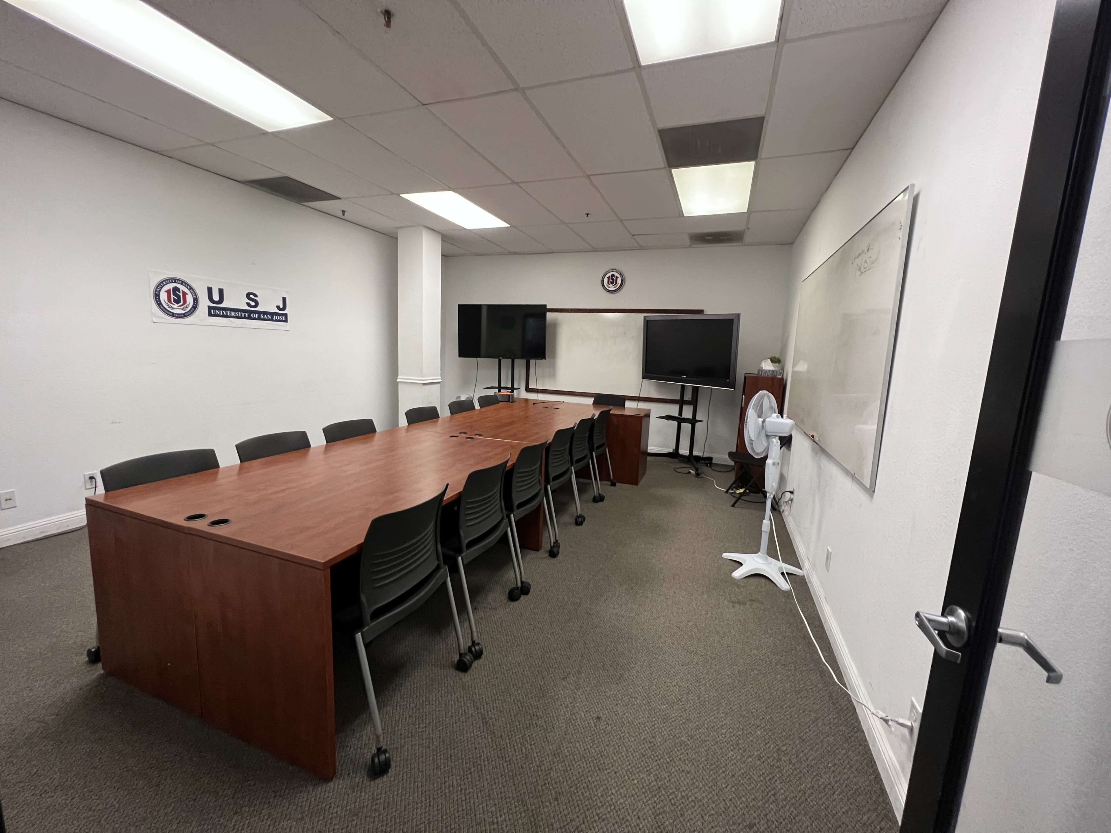 A conference room with a long wooden table surrounded by gray chairs, multiple screens mounted on the wall, and a whiteboard.