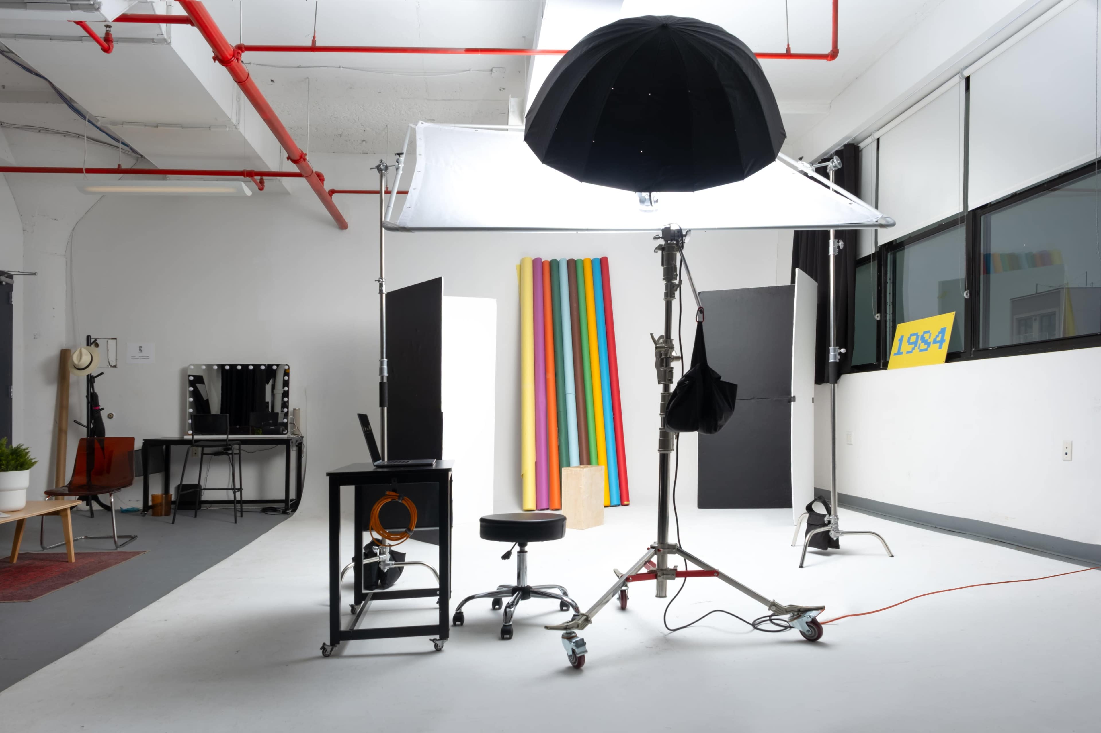 The image shows a photography studio setup with a large umbrella light, a backdrop of colorful stripes, and various equipment including a stool and a mirror.