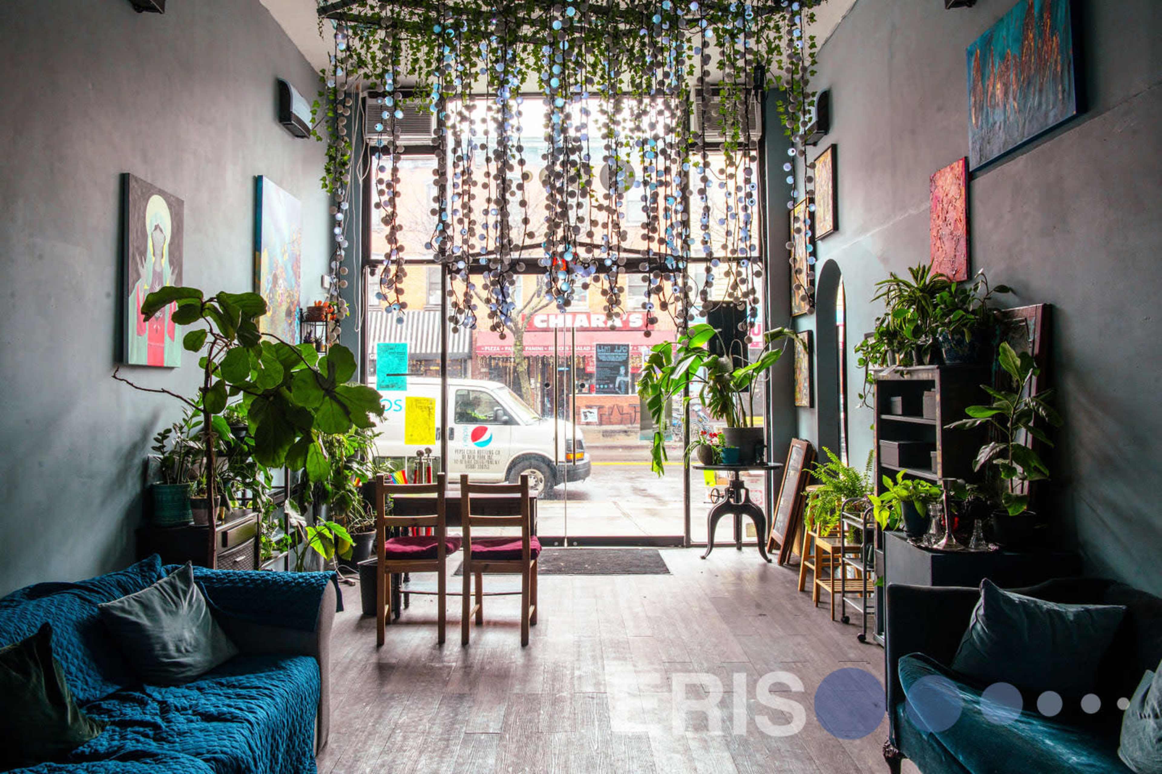 A cozy cafe interior filled with plants, featuring wooden furniture and a large window letting in natural light.