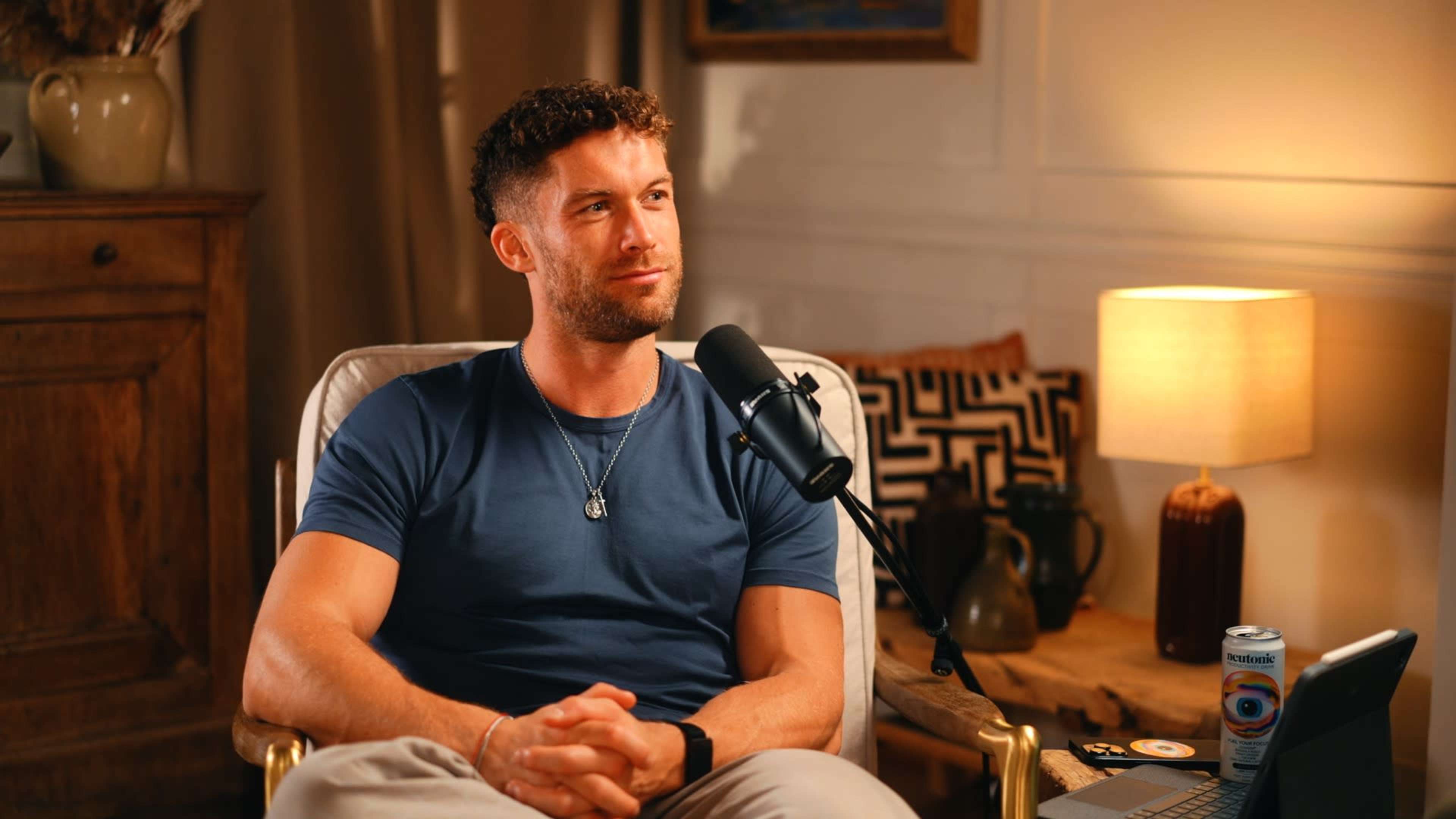 A man with curly hair sits in a chair in a well-lit room, facing a microphone and a laptop, with a lamp and decorative items in the background.