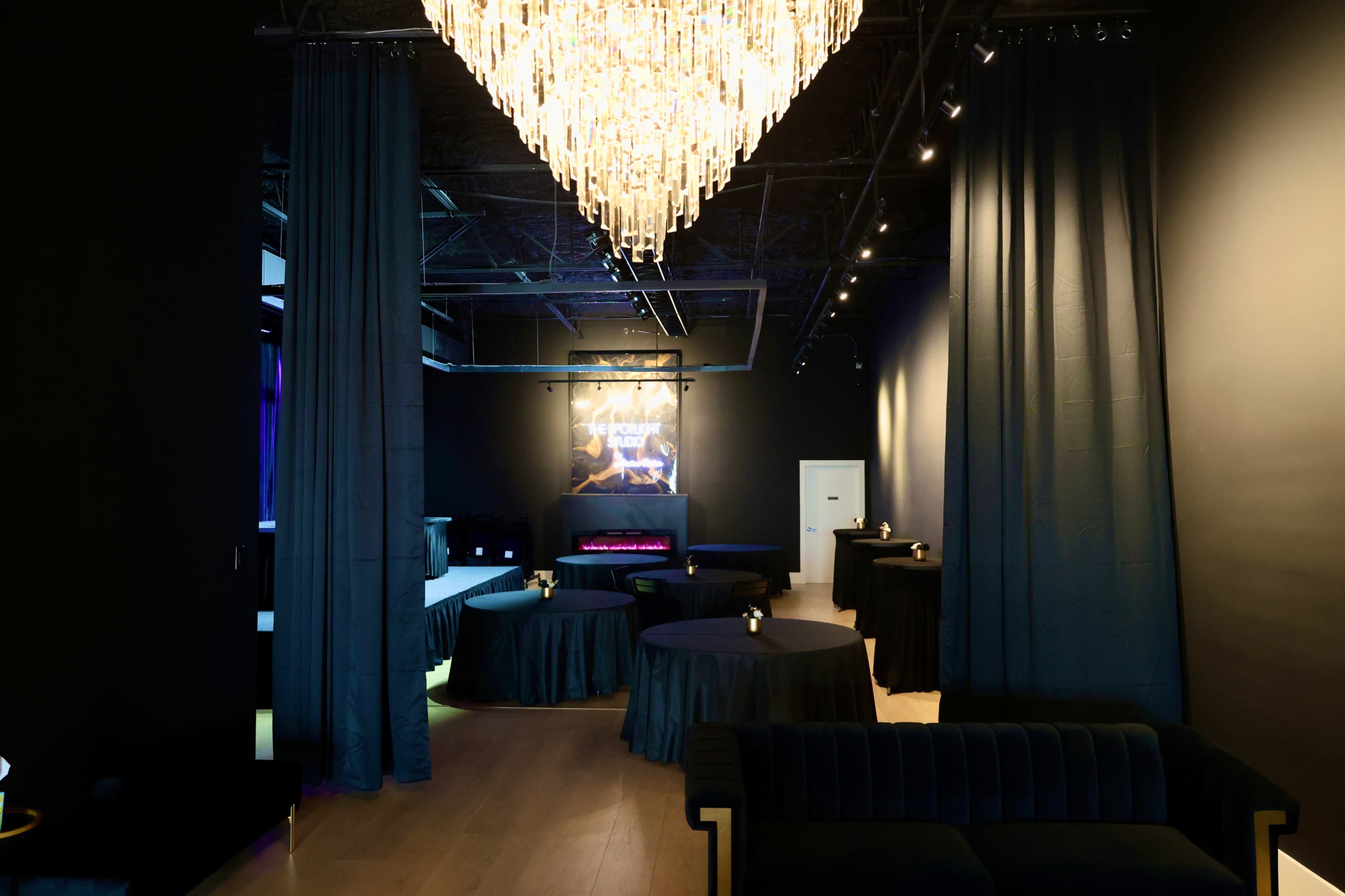 The image shows an elegantly designed interior space featuring dark drapes, circular tables with tablecloths, and a chandelier hanging from the ceiling.