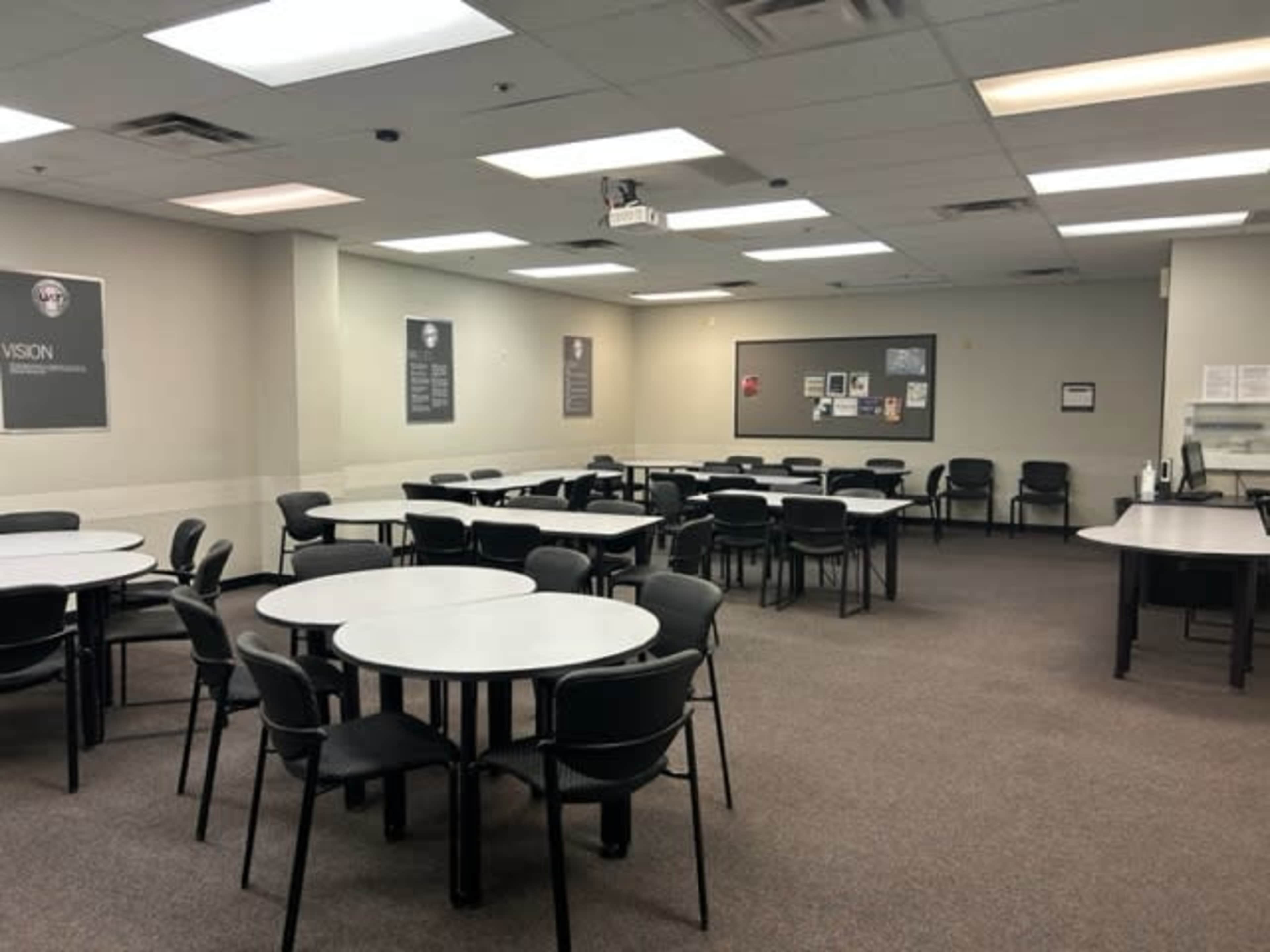 A spacious room with multiple round tables and chairs arranged neatly, along with a blackboard and a wall displaying various posters.