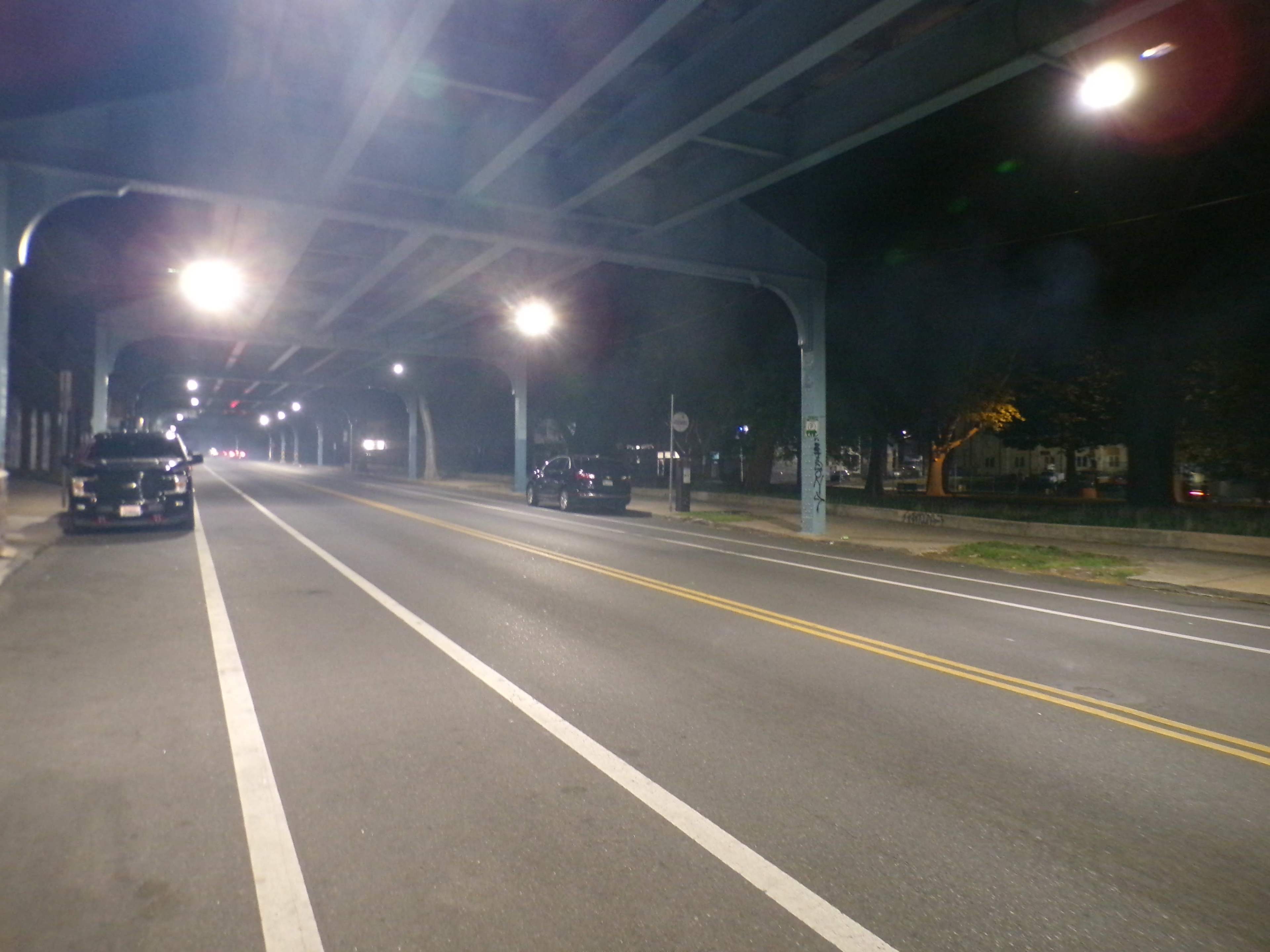 The image shows a dark, empty street illuminated by overhead lights, with a few vehicles parked along the side under a metal overpass.